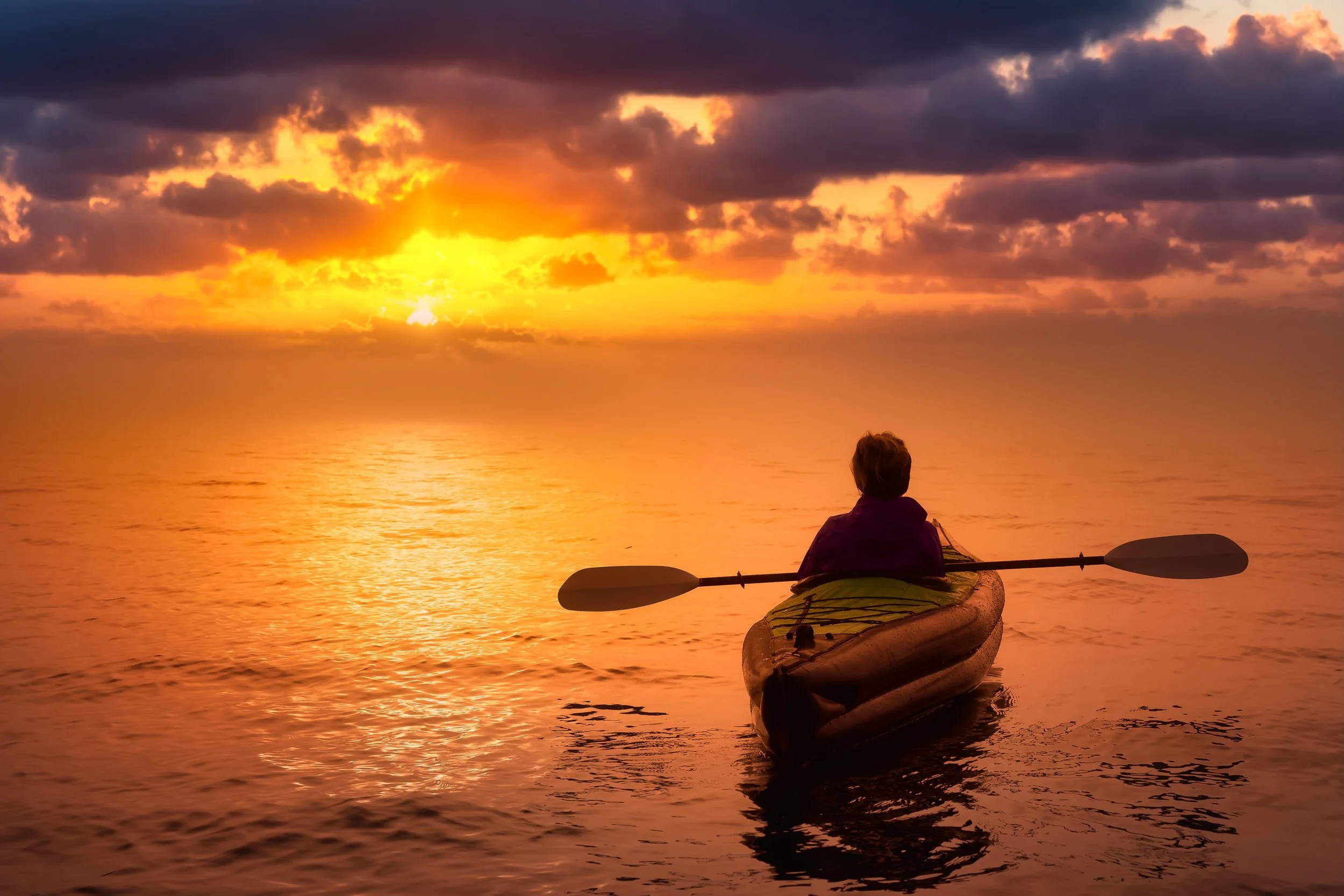 Person kayaking on the water during a colorful sunset with clouds in the sky.