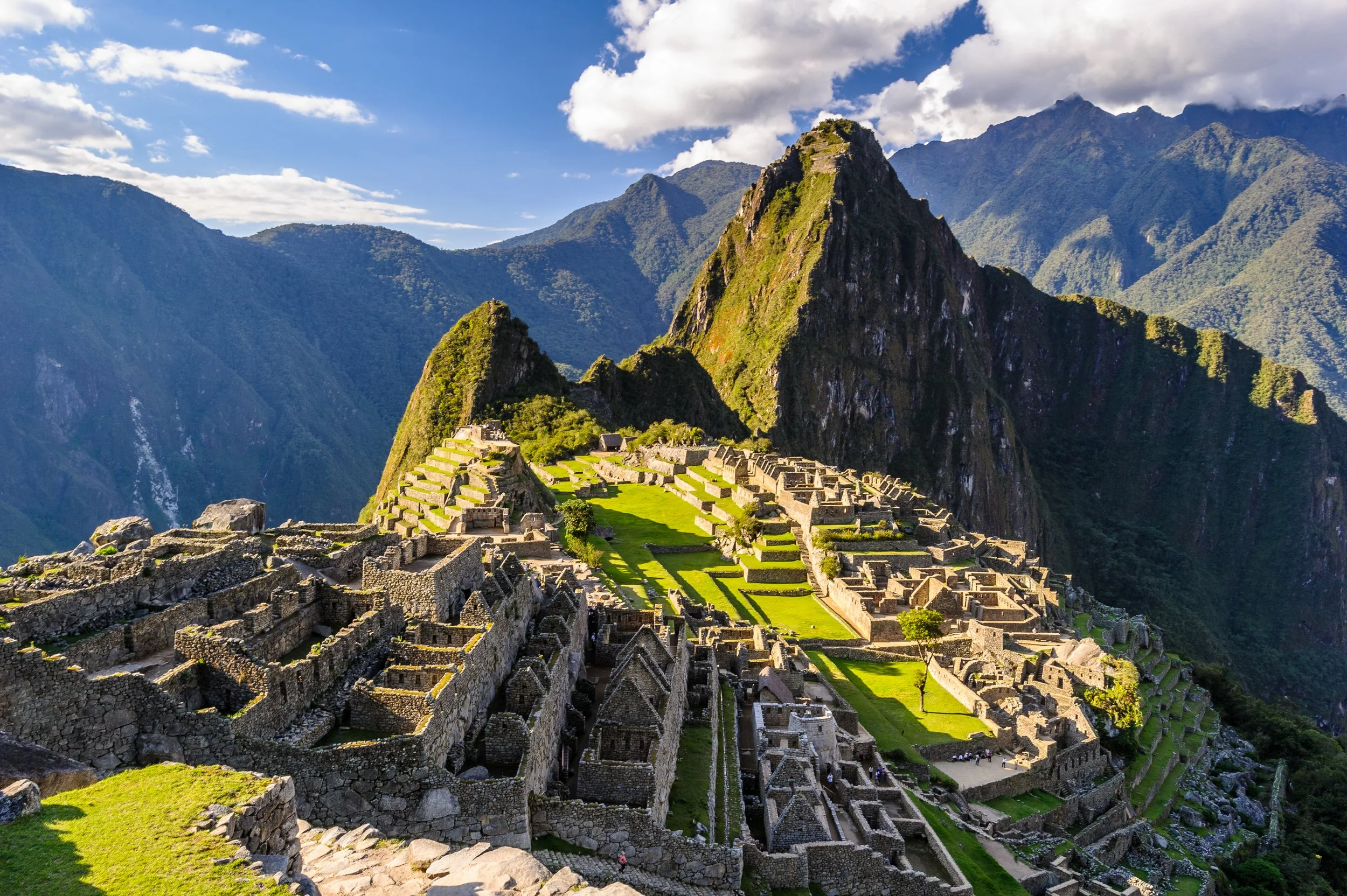 Machu Picchu ruins with stone structures and terraces, surrounded by lush green mountains under a partly cloudy sky.