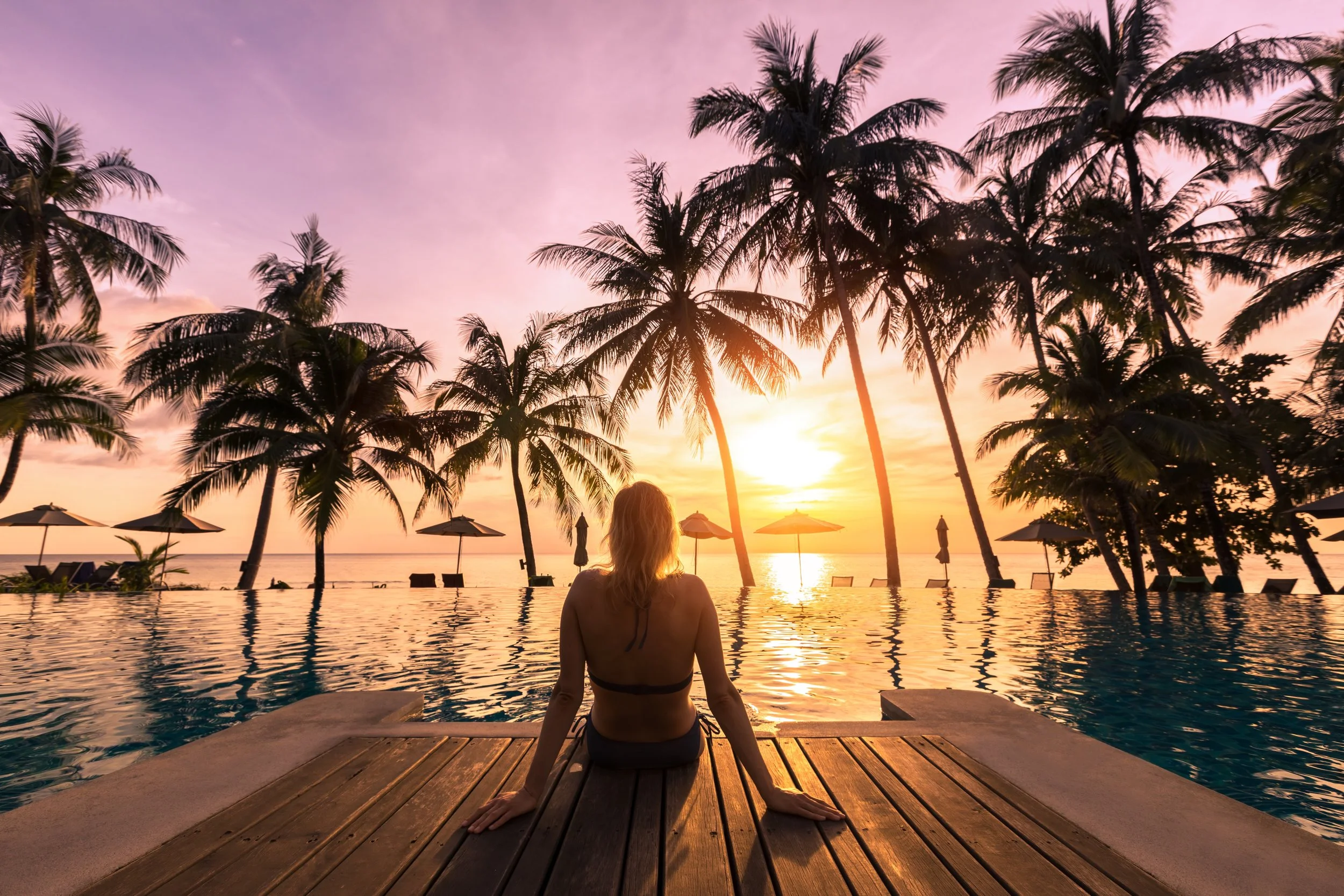 Woman sitting by a swimming pool at sunset, surrounded by palm trees and beach umbrellas, with the ocean in the background
