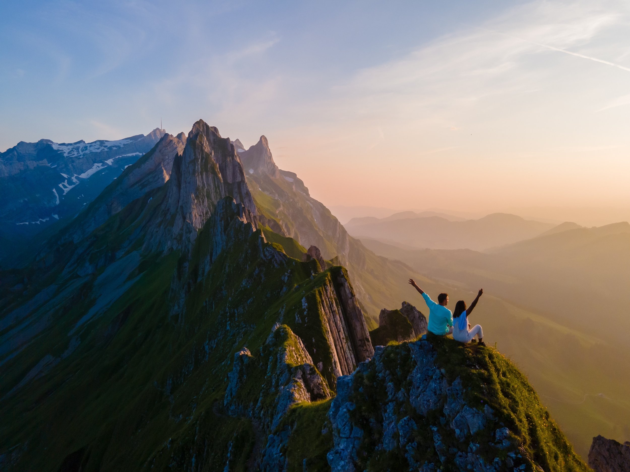 A couple sitting on a mountain summit at sunset with arms raised, overlooking a scenic mountain range with green slopes and snow-capped peaks.
