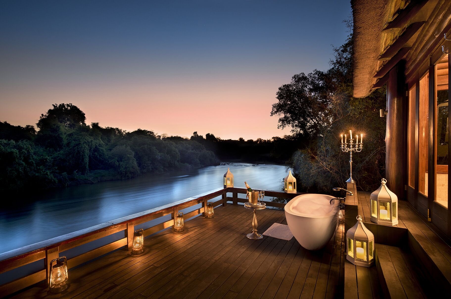 An outdoor deck with a bathtub, lanterns, a candelabra, and a view of a river at dusk, with trees on the riverbank and a clear sky.