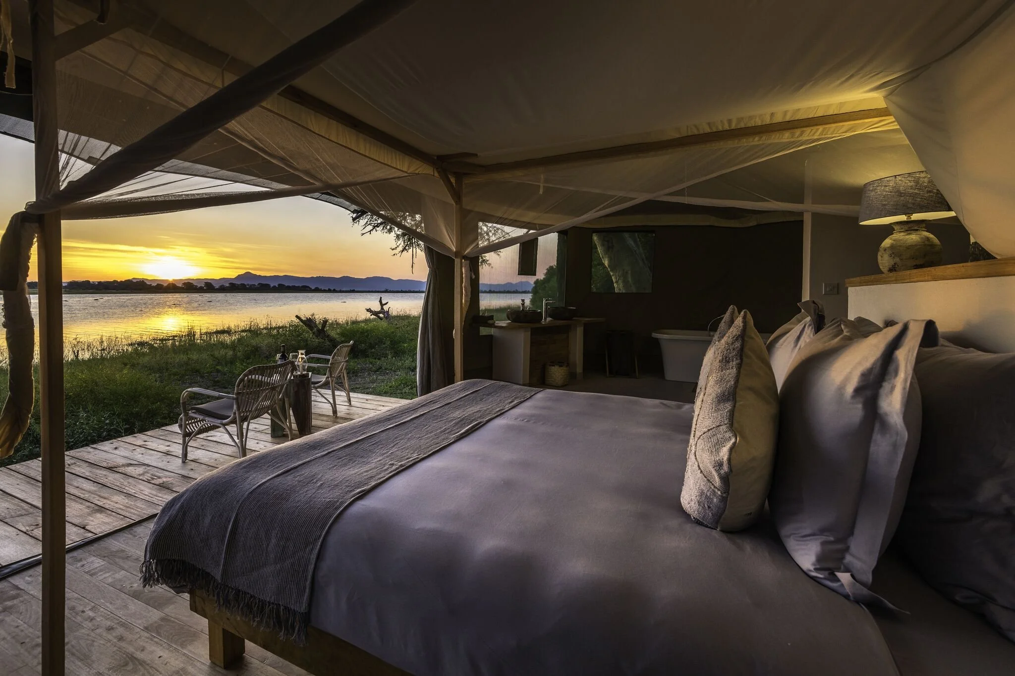 Inside a cozy, luxurious tented accommodation overlooking a body of water at sunset, with a large bed, pillows, a bath in the background, and a sitting area outside on the deck.