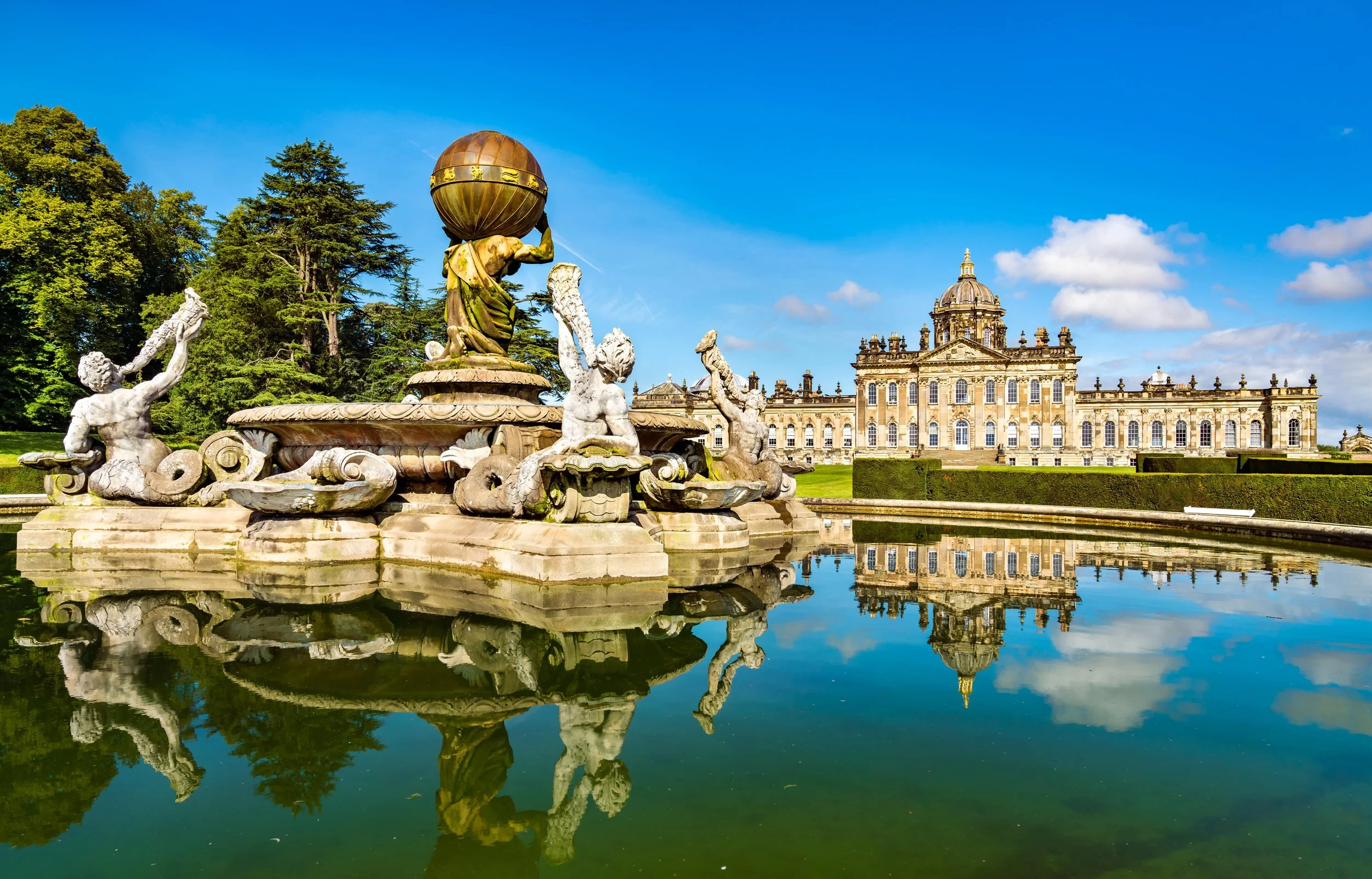 Baroque-style fountain with sculptures of mythological figures and a large orb, reflected in a pool, in front of a historic building with classical architecture, under a blue sky with clouds.