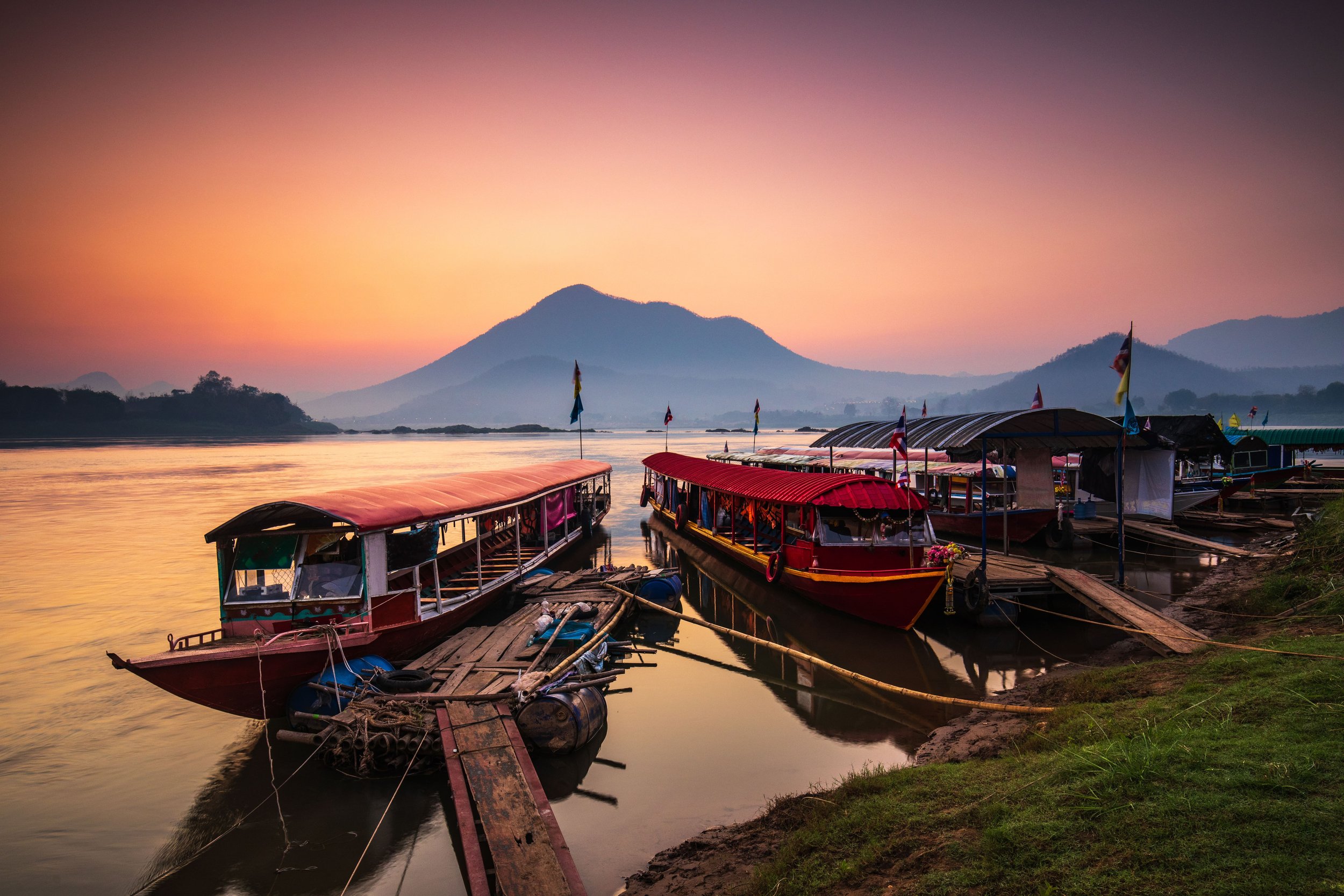 Boats docked along a river at sunset with mountains in the background.