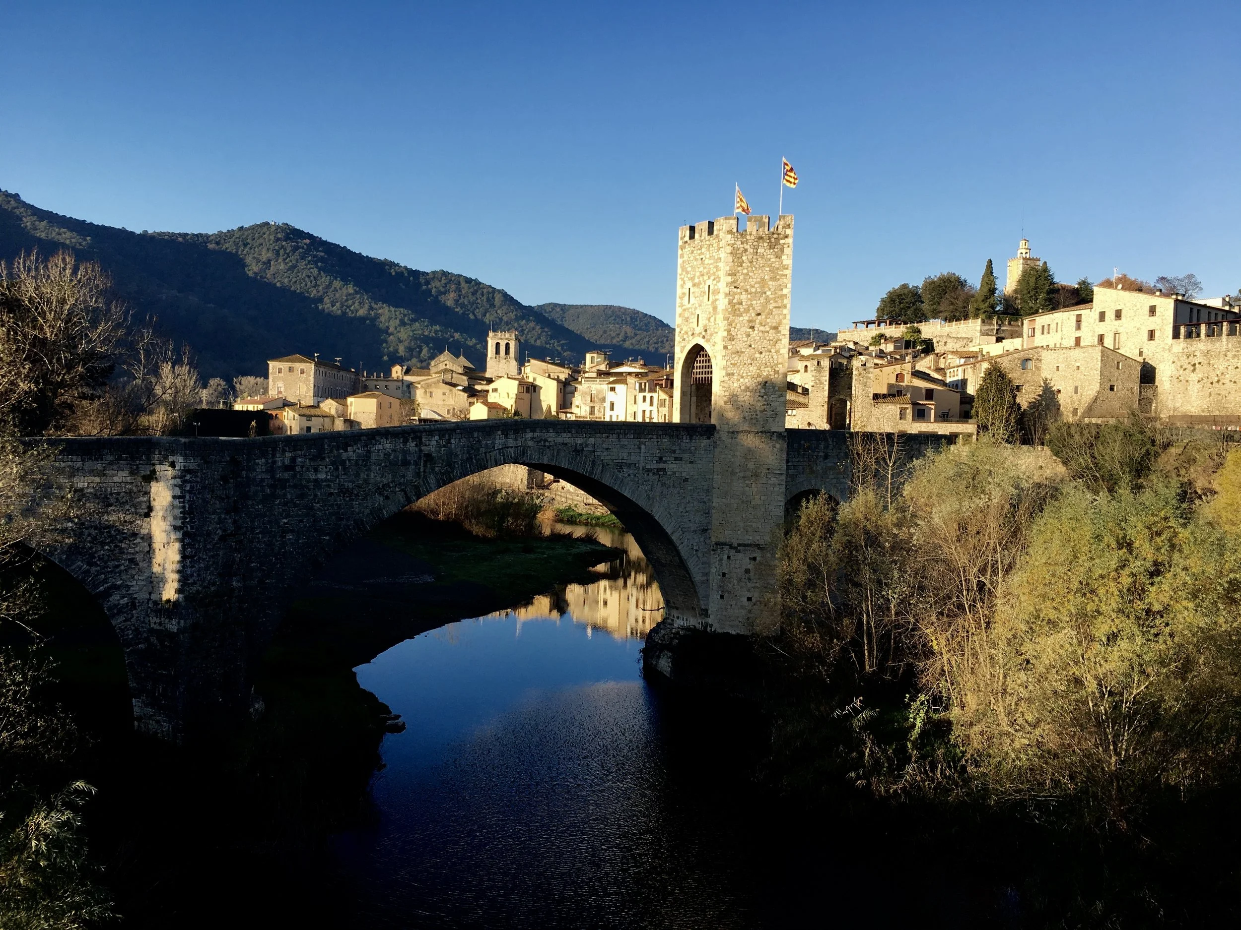 Historic stone bridge over a river with a nearby medieval tower and a village with stone buildings on a hillside, mountain background, clear blue sky.