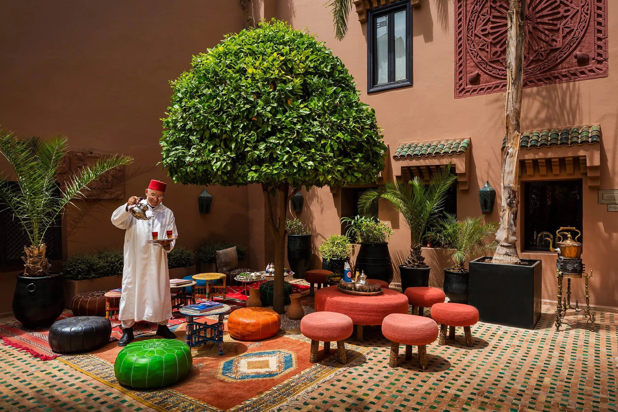 Man dressed in traditional Moroccan attire pouring tea in a Moroccan-style courtyard with large potted plants, colorful poufs, and a central tree.