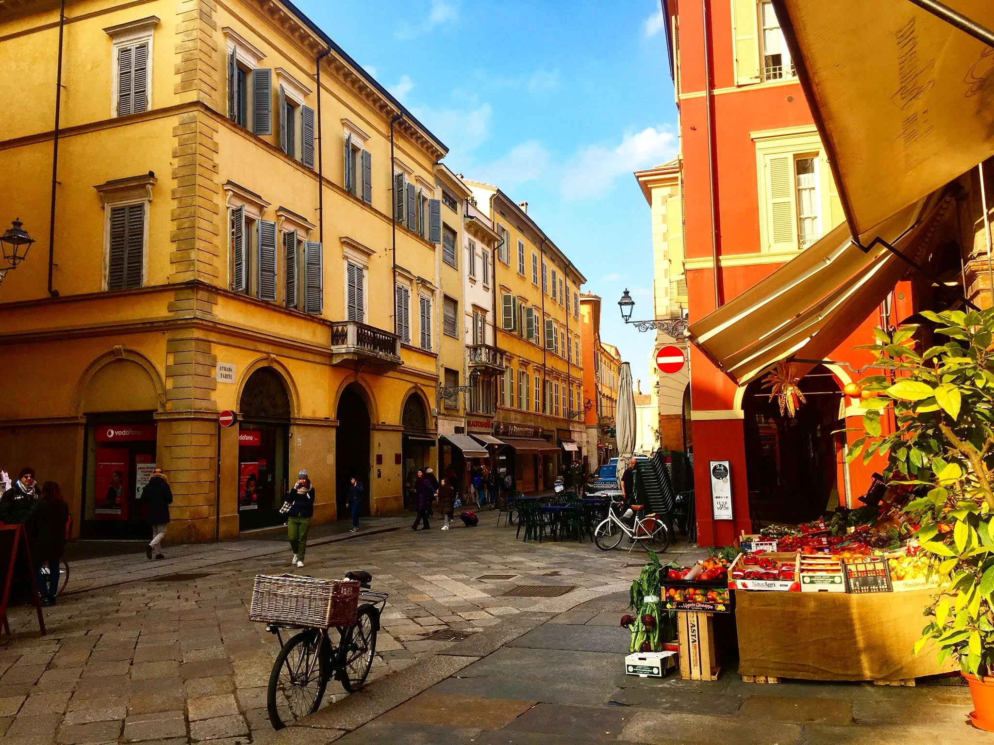 A street scene in a European city with colorful buildings, pedestrians, a bicycle, and an outdoor fruit stand.