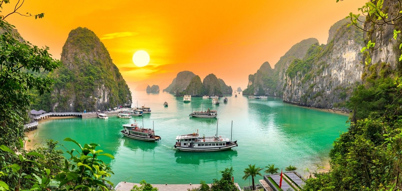 A scenic harbor at sunset with multiple boats floating on greenish-blue water, surrounded by lush green forested cliffs and islands.