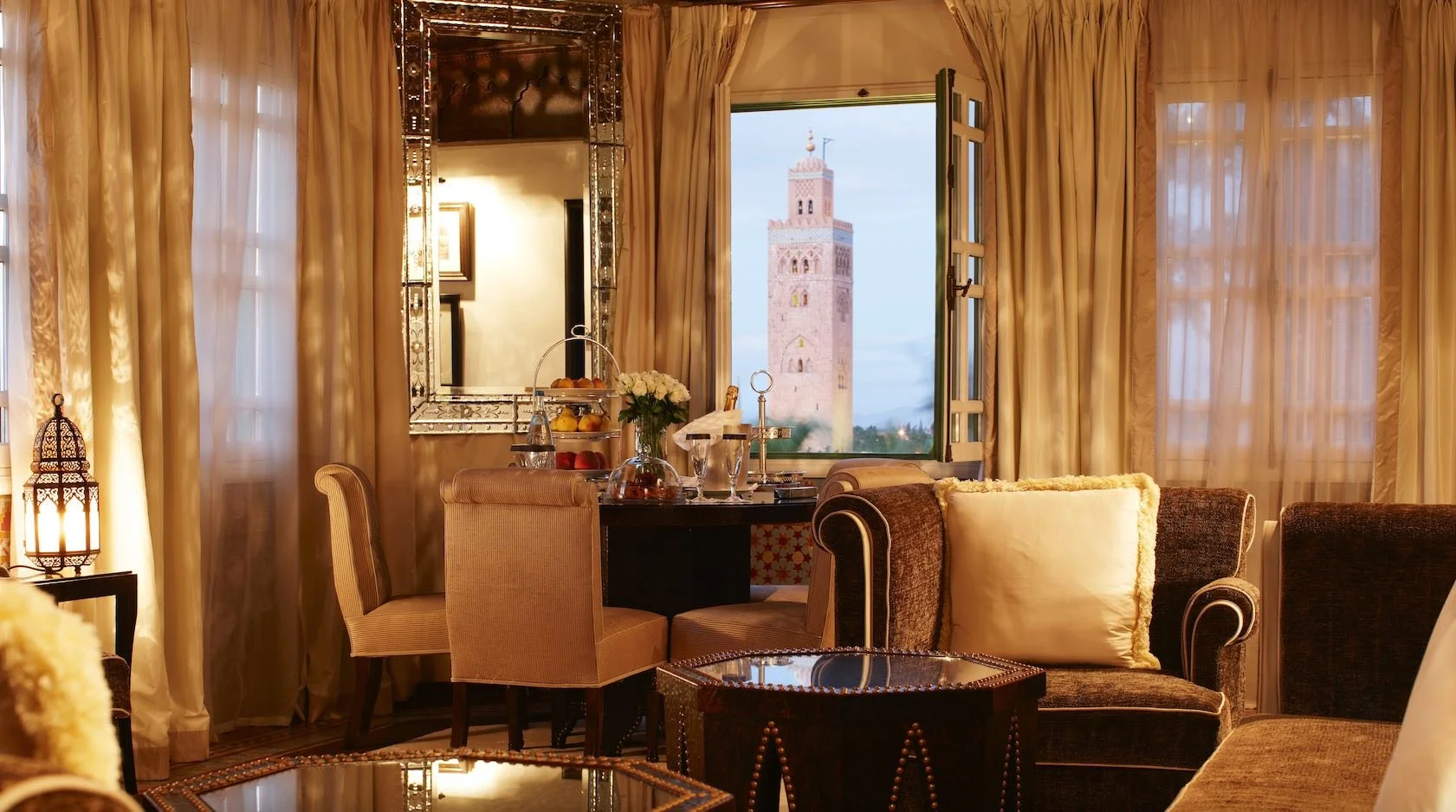 A cozy living room with beige curtains, a sofa with a large cream pillow, a round glass-top coffee table, a table with flowers, drinks, and snacks, and an open window showing a tall tower against a sky during dusk.