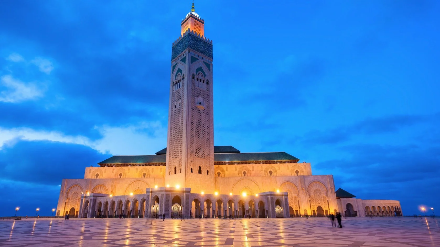 The Hassan II Mosque in Casablanca, Morocco, illuminated at dusk with a blue sky and clouds in the background, featuring a large patterned plaza in front.