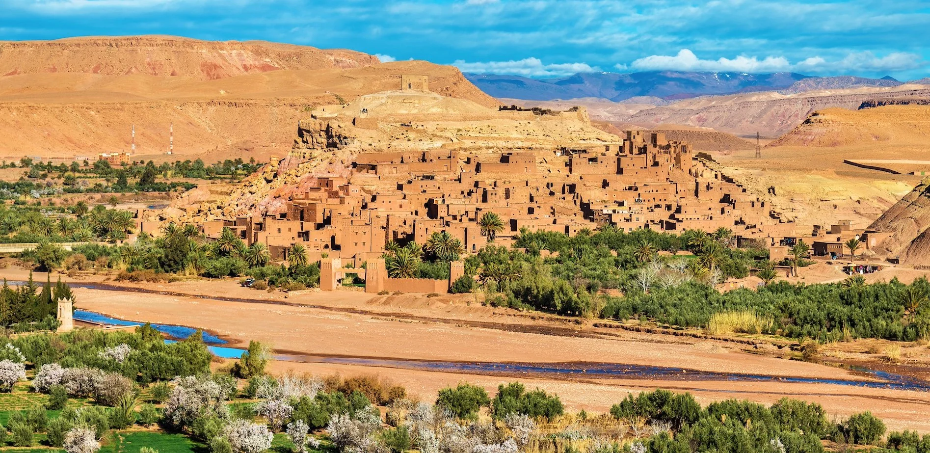 A desert town built into a hillside with terraced adobe-style buildings, surrounded by palm trees and greenery, with a river in the foreground and mountains in the background under a blue sky.