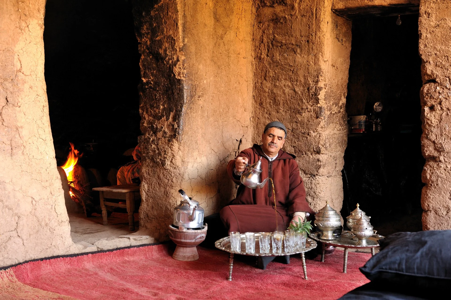 A man dressed in traditional Moroccan clothing sitting on a red rug in a cave-like space, pouring tea from a silver teapot into small glasses placed on a tray in front of him, with metallic containers and a kettle nearby.