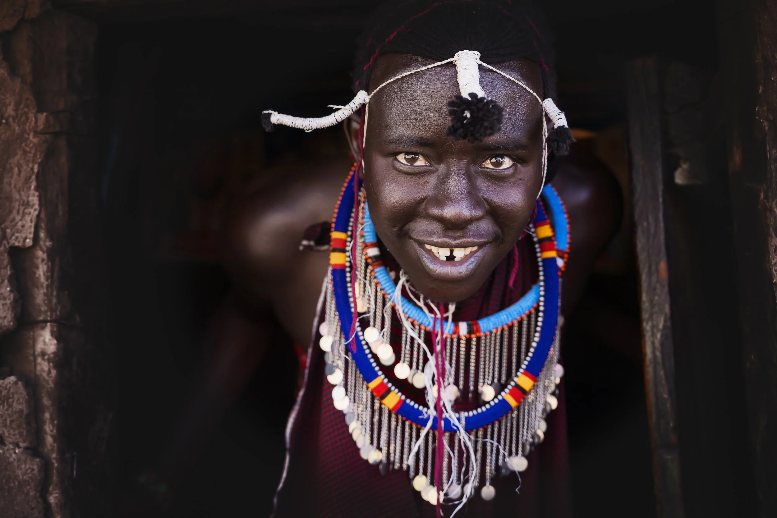 A smiling woman from the Maasai tribe wearing traditional beaded jewelry, crawling out of a dark wooden structure.