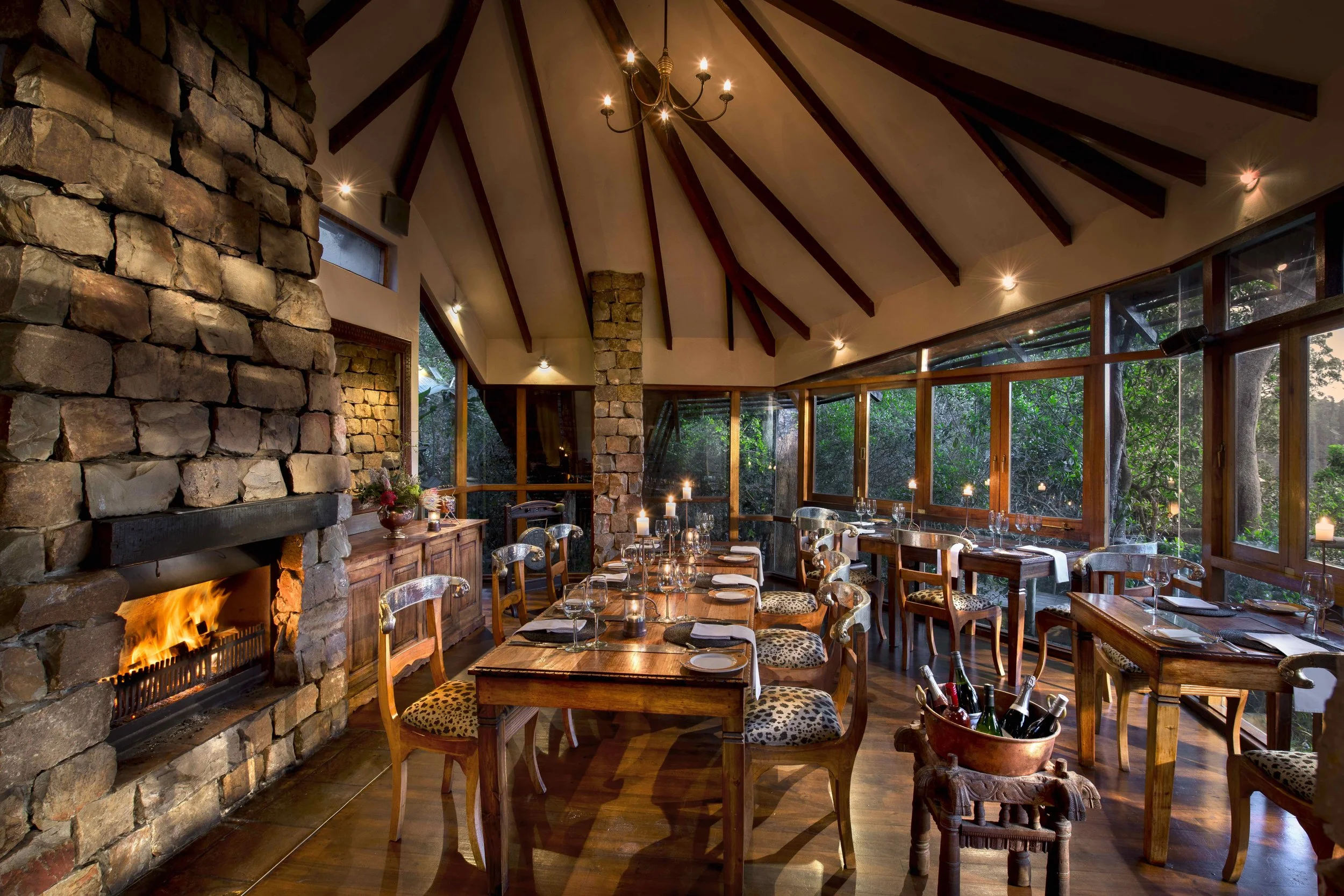 A cozy restaurant interior with a stone fireplace, wooden furniture, and large windows overlooking greenery, decorated for evening dining.