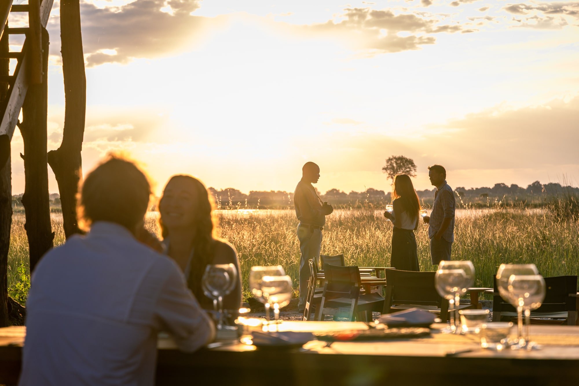 People dining at an outdoor restaurant during sunset, with four individuals standing and talking in a field background and two people seated in the foreground with glasses and tableware.