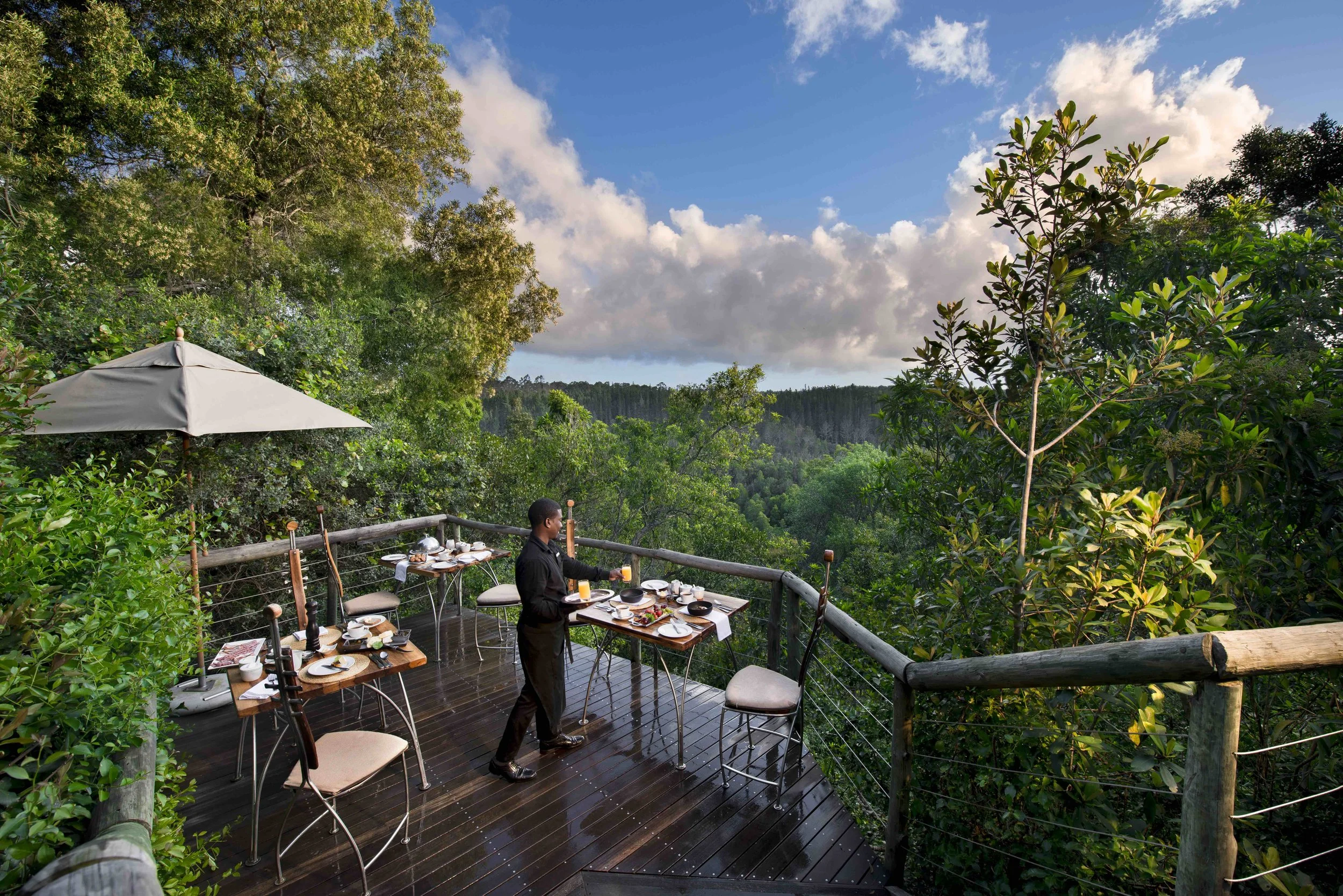 A dining setup on a wooden deck overlooking lush green forest and cloudy sky, with a waiter setting the table.