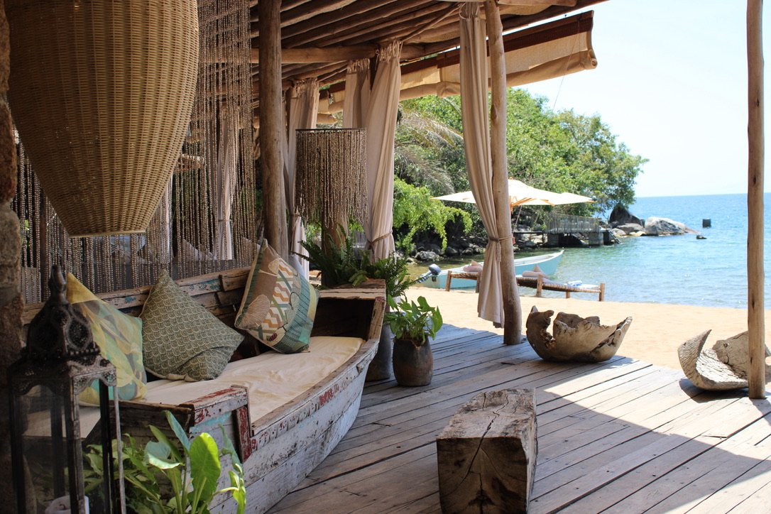 A rustic outdoor beachside lounge area with a wooden sofa, pillows, plants, and hanging curtains, with the sandy beach, ocean, and boats in the background.