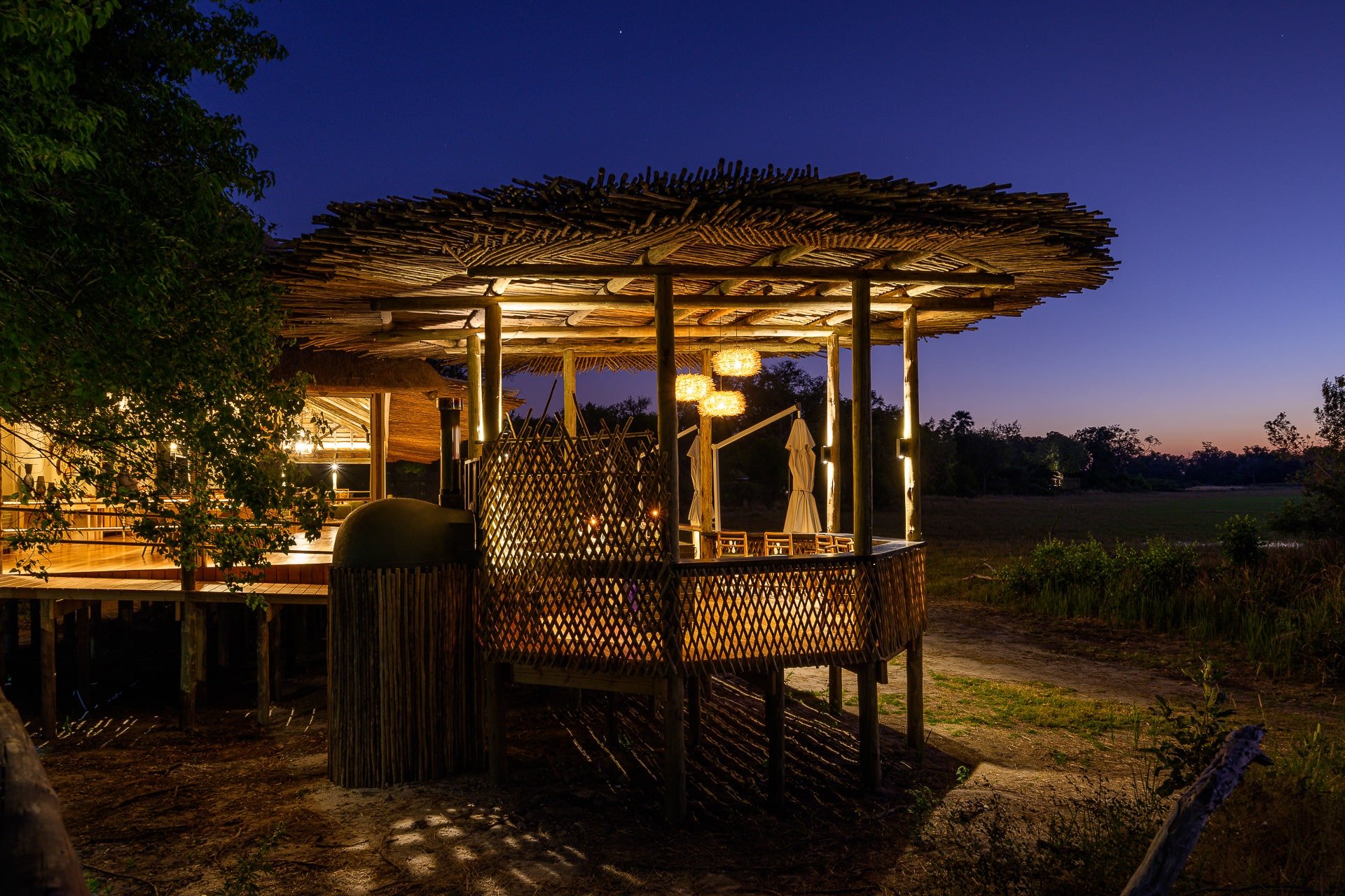 A wooden outdoor restaurant or bar with a thatched roof illuminated by warm lights, set against a twilight sky with trees and open land in the background.