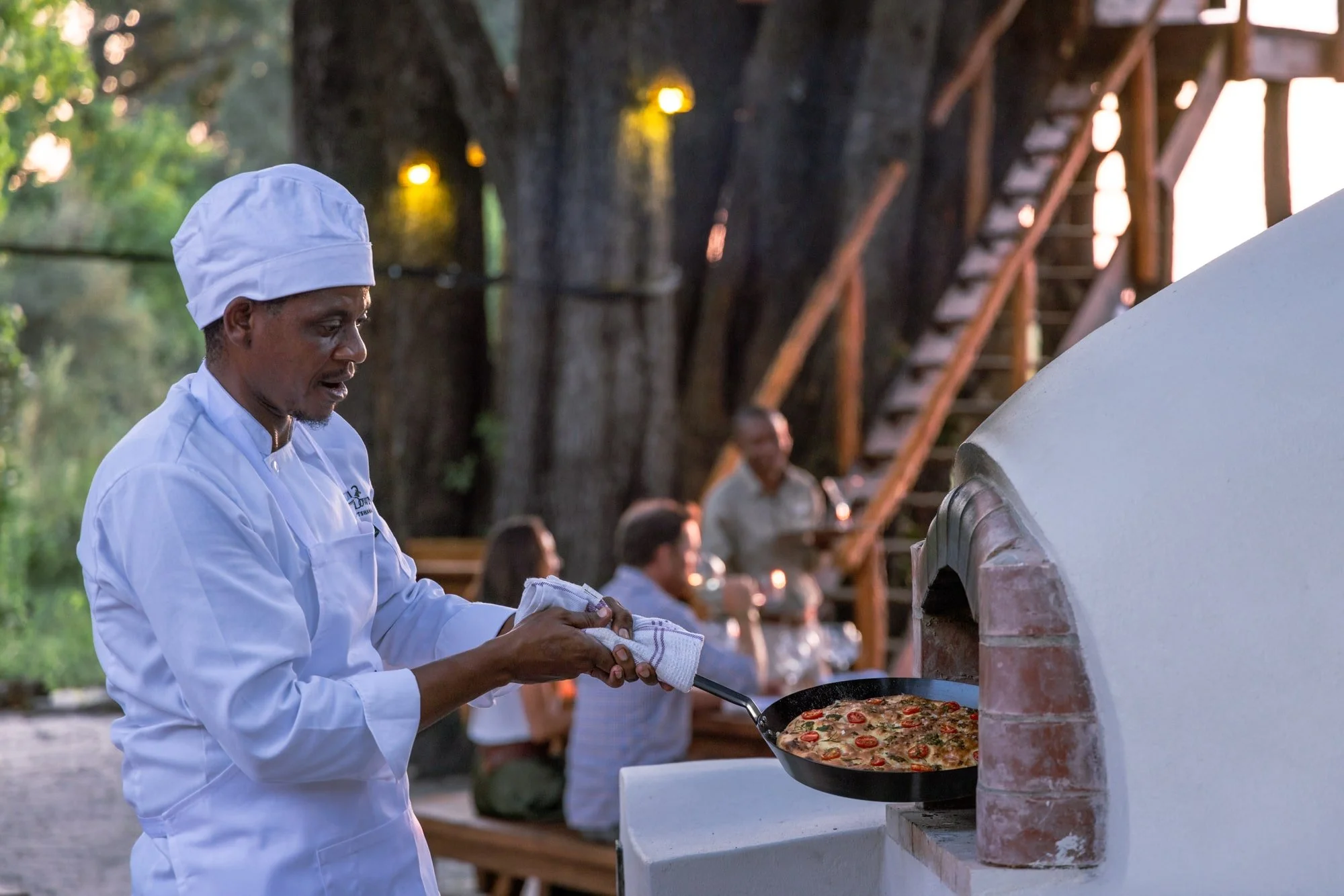 A chef in white clothing and a hat is cooking pizza in an outdoor stone oven. In the background, people are sitting at a wooden table with a tree and string lights nearby.