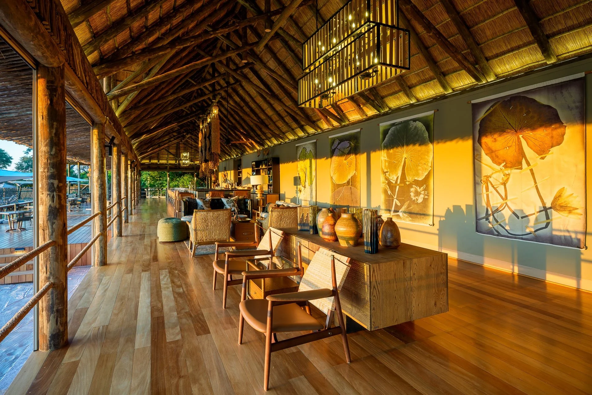 Indoor dining area with a thatched roof ceiling, wooden flooring, modern chairs, a sideboard with decorative vases, illuminated by warm sunset light, large framed leaf artwork on the wall, and outdoor seating visible through the open side.