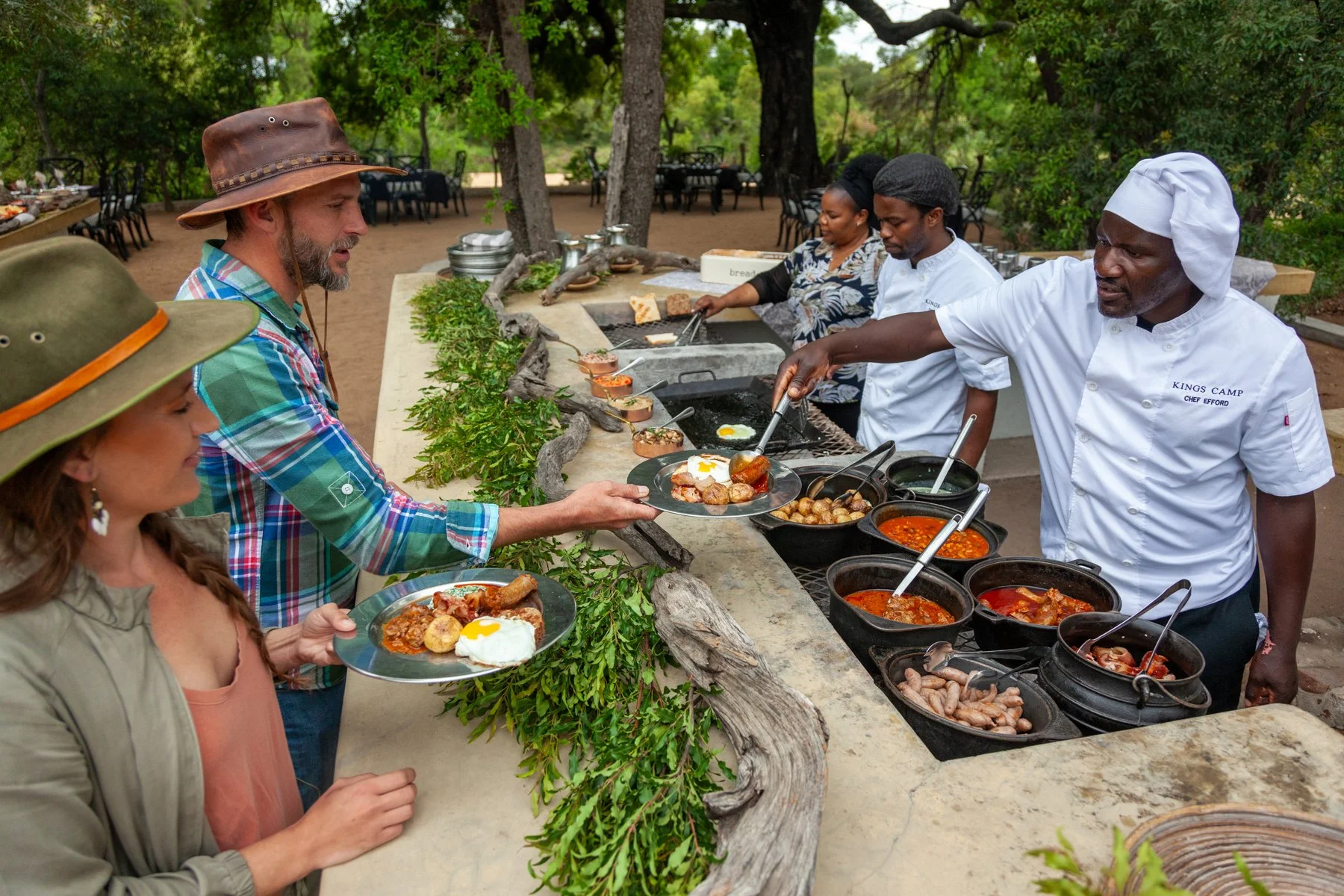 People in an outdoor setting at a buffet-style breakfast with various dishes, chefs serving food, and a person receiving a plate of food.
