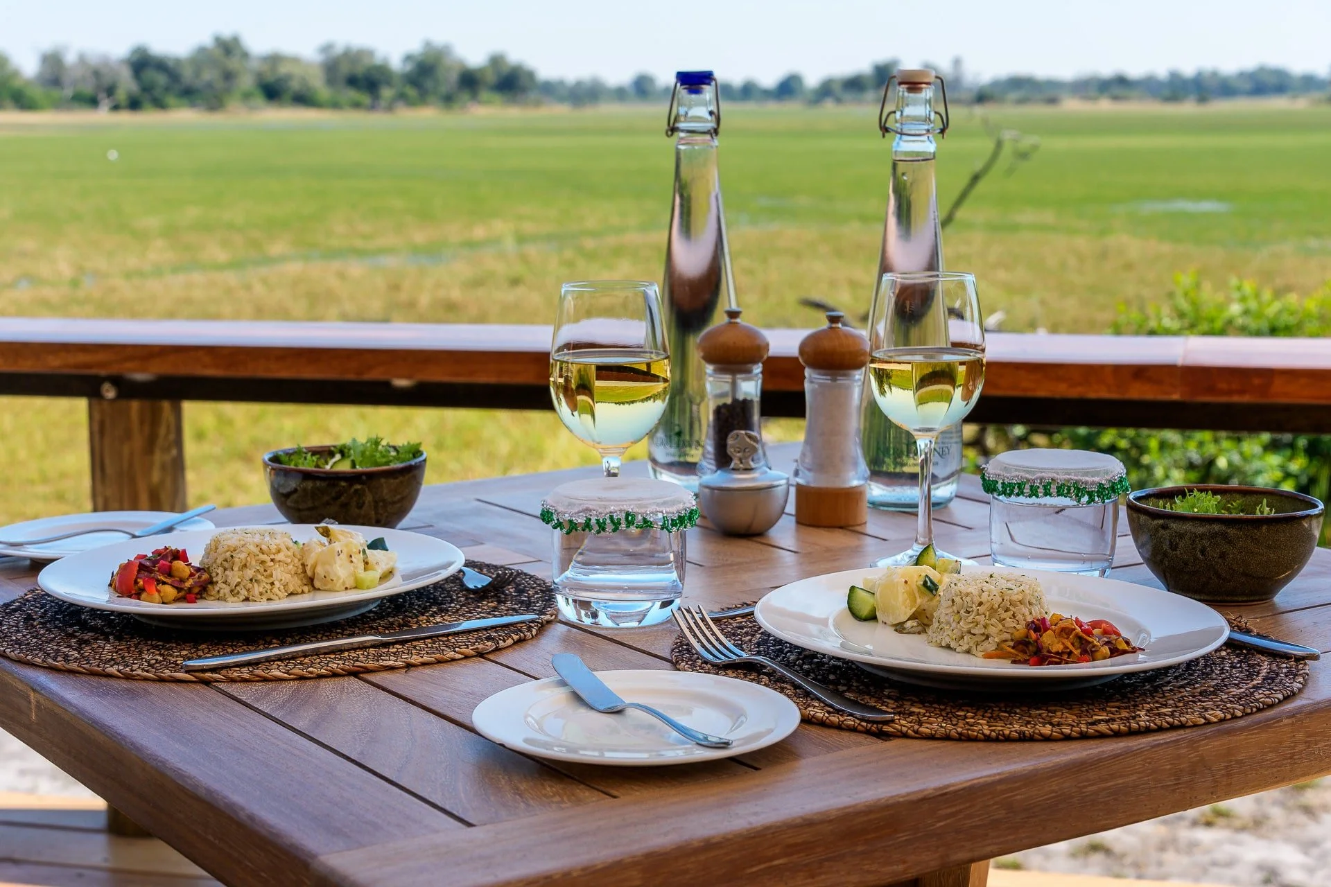 Table set outdoors with two plates of rice, vegetables, and potatoes, two glasses of white wine, water, bowls of salad, salt and pepper shakers, water bottles, and a garnish, overlooking a green field.