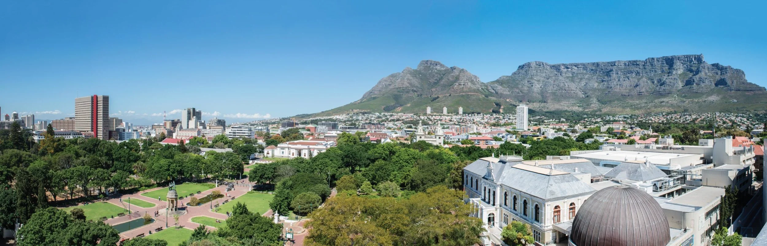 Panoramic view of Cape Town city with Table Mountain in the background, green park in the foreground, and various buildings and skyscrapers under a clear blue sky.