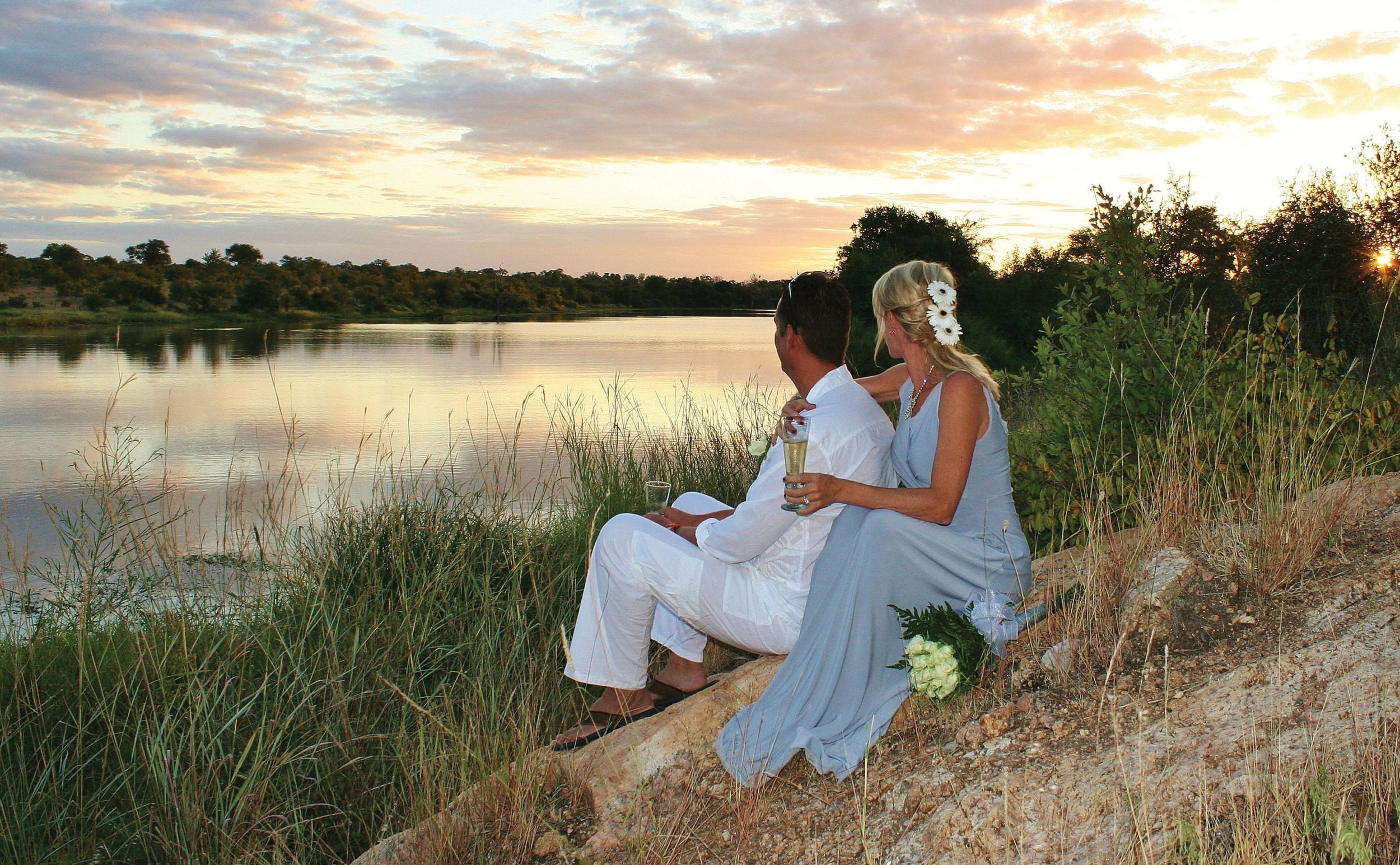 A man and woman sitting on a rock by a river at sunset. The woman has flowers in her hair and is holding a glass of champagne, while the man is also holding a glass of champagne. They are dressed in light-colored clothing and look at the river and su
