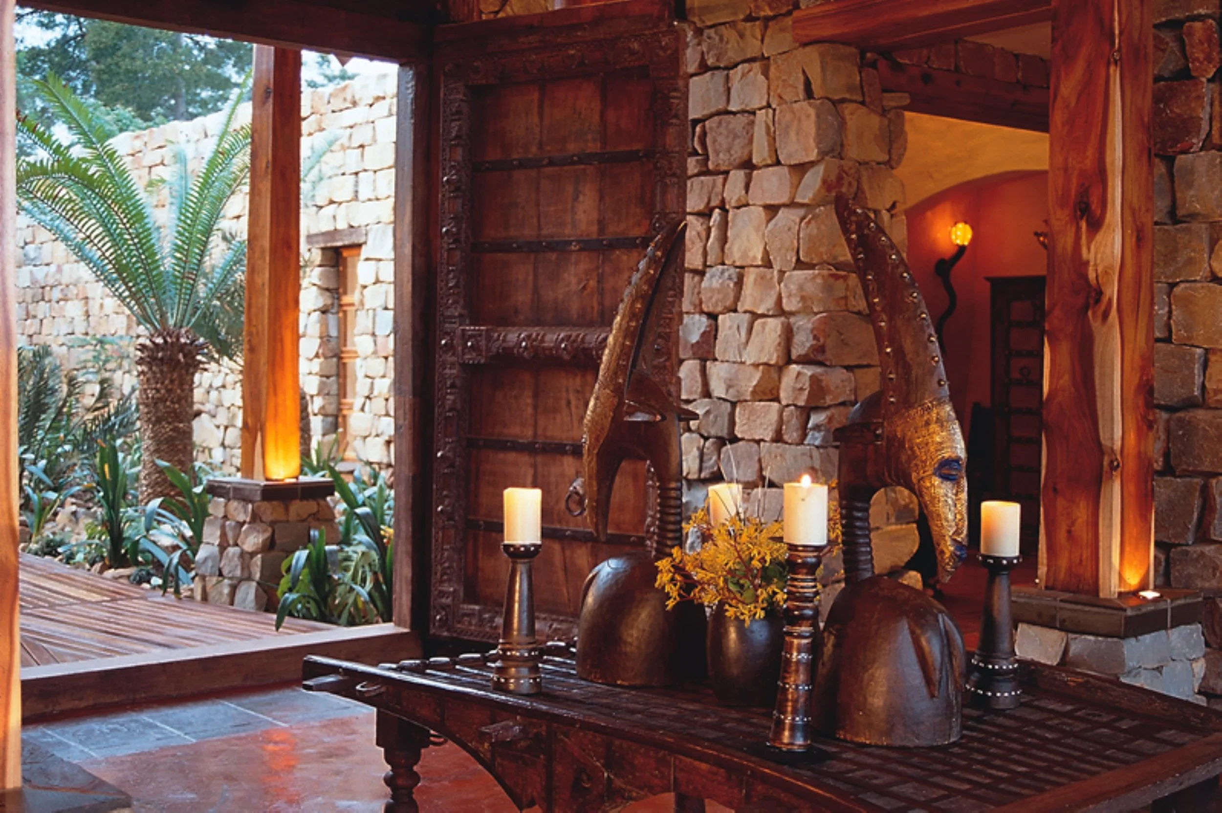 Interior of a rustic-style room with stone walls, wooden accents, and African tribal masks on a wooden table, decorated with candles and yellow flowers, with an outdoor view of lush greenery and palm trees through an open doorway.