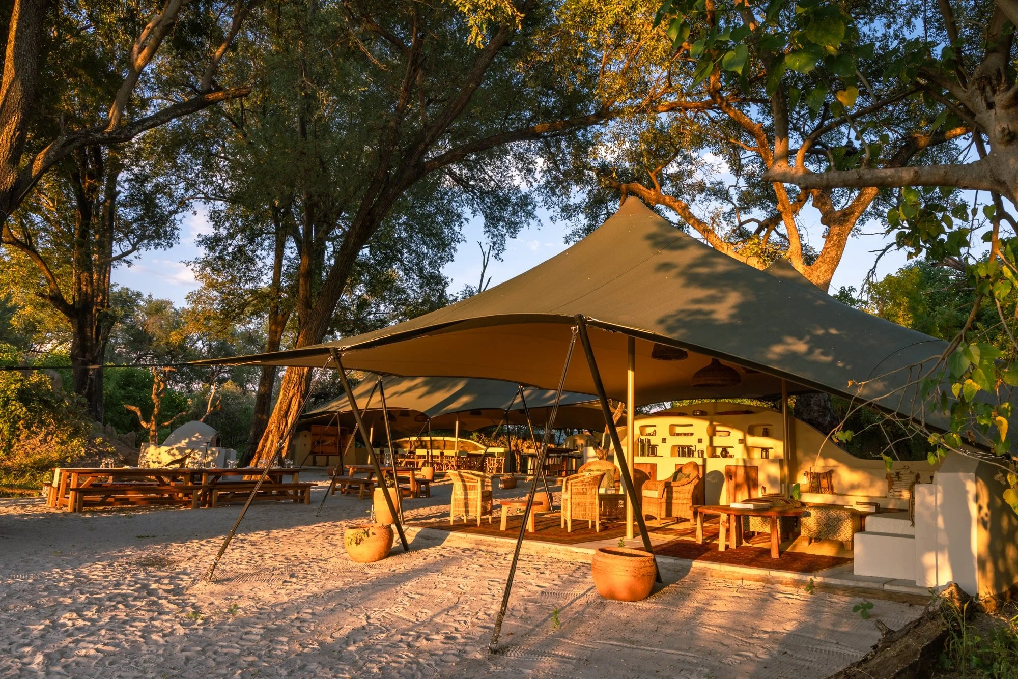 Outdoor lounge area with large canopy tent, wooden furniture, and sand ground, surrounded by trees at sunset.