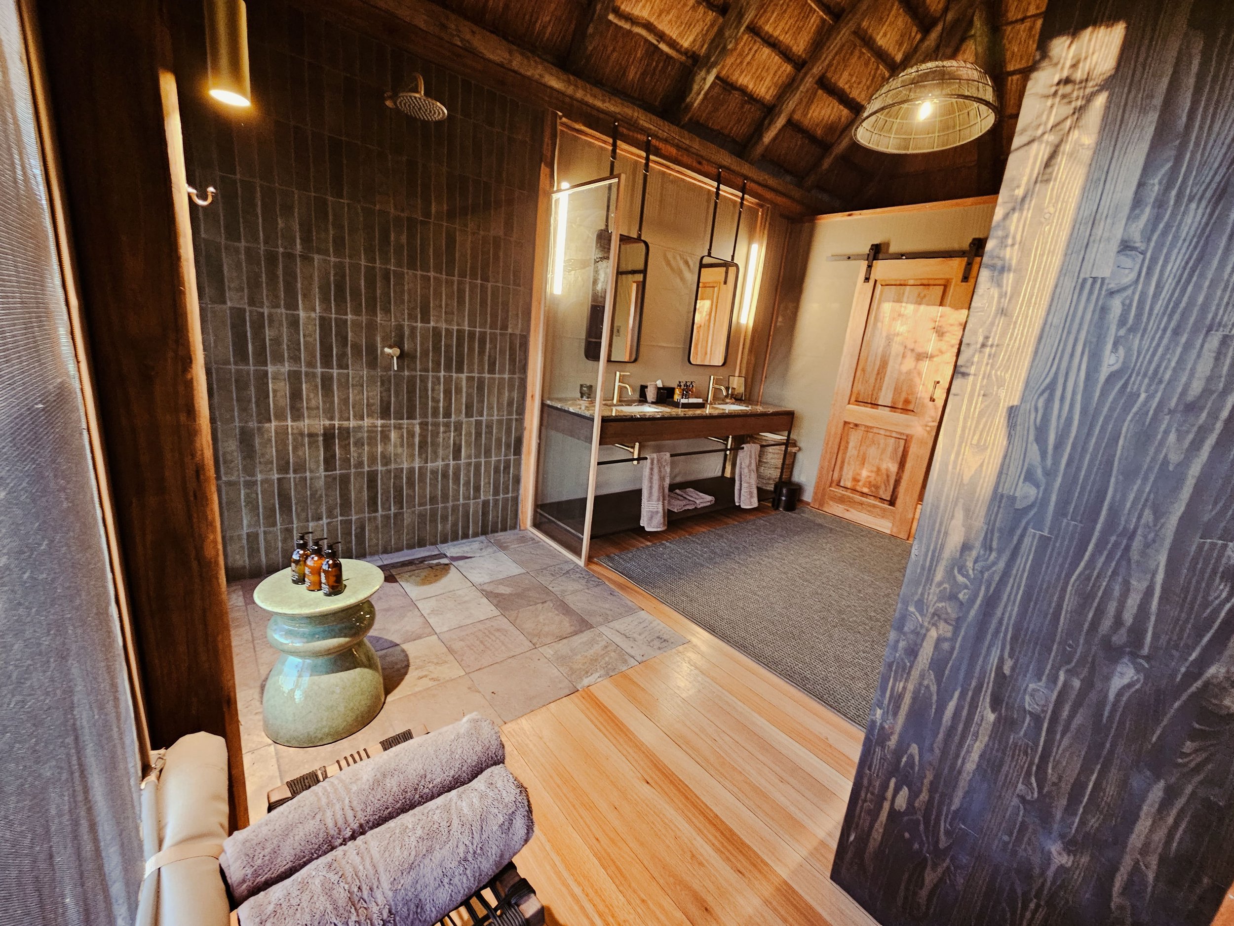 A rustic bathroom featuring a walk-in shower with dark brown tiles, a double sink vanity with mirrors, and wooden accents on the doors and ceiling.