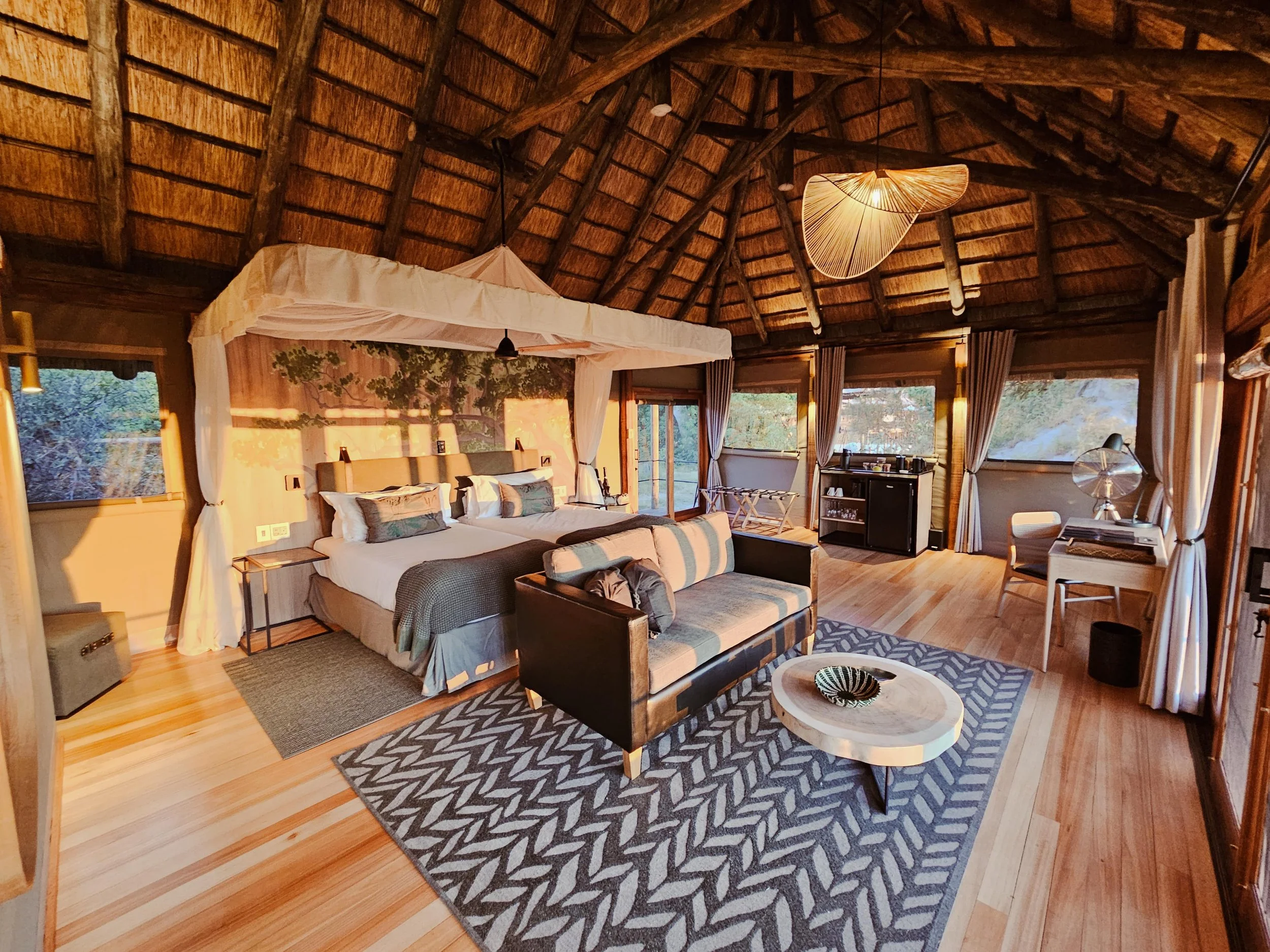 Luxury hotel room with a thatched ceiling, large bed with white canopy, sofa, coffee table, and various furniture, surrounded by windows with curtains and warm lighting.