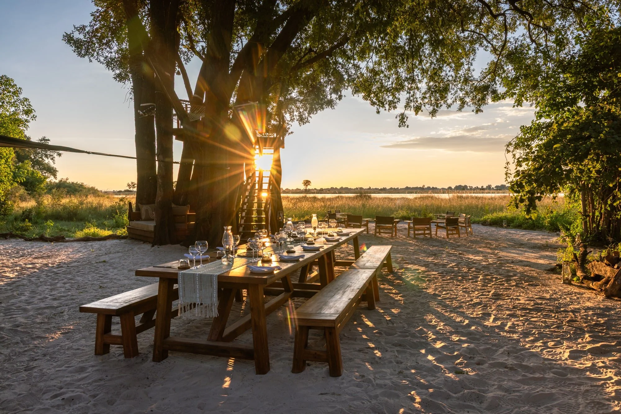 Outdoor dining setup with long wooden table, benches, wine glasses, and a table runner, situated on sandy ground beneath large trees at sunset with a view of a grassy field and water in the background.