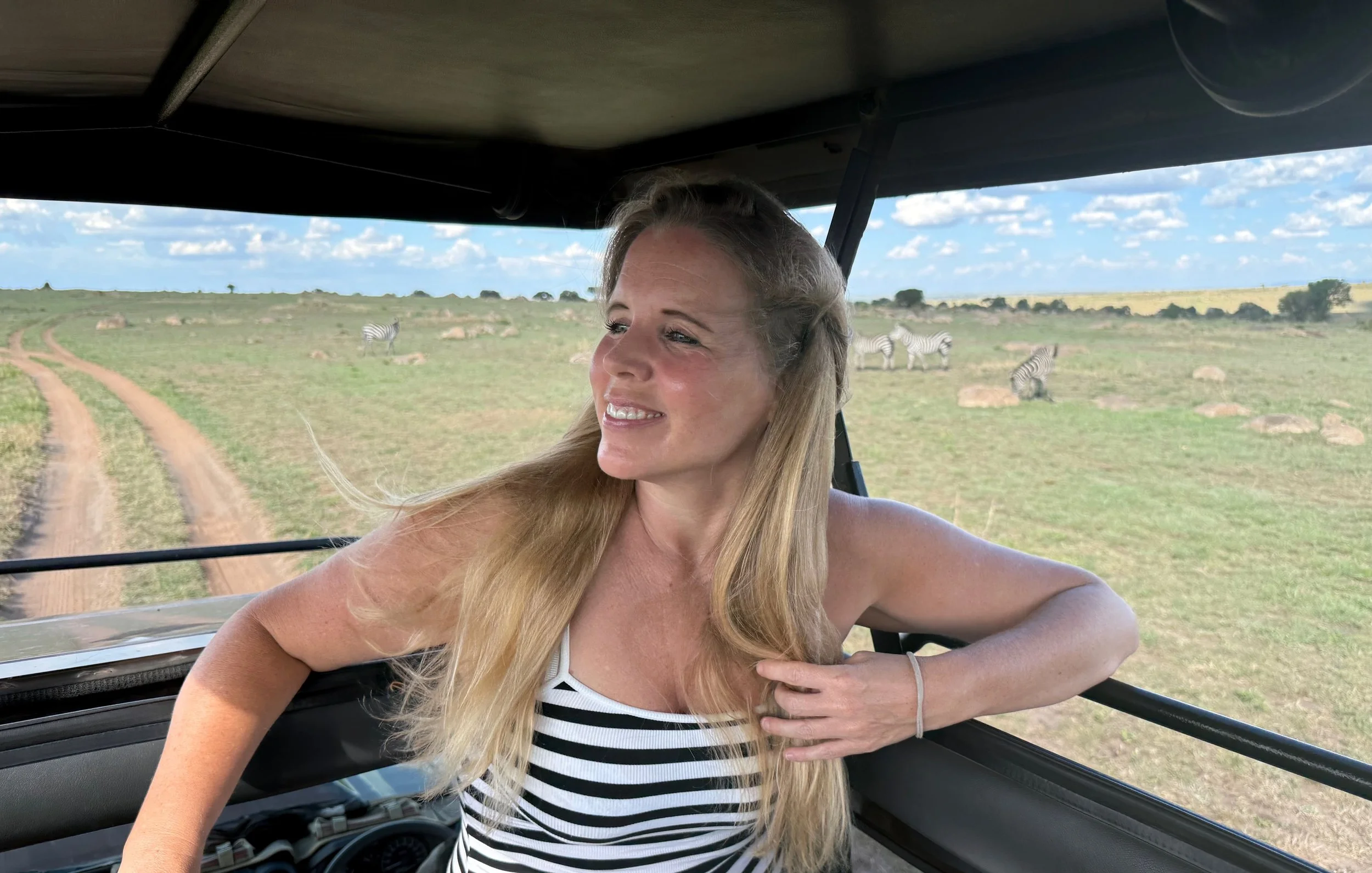 Woman with long blonde hair wearing a black and white striped tank top inside a safari vehicle with a view of zebras and open landscape in the background.