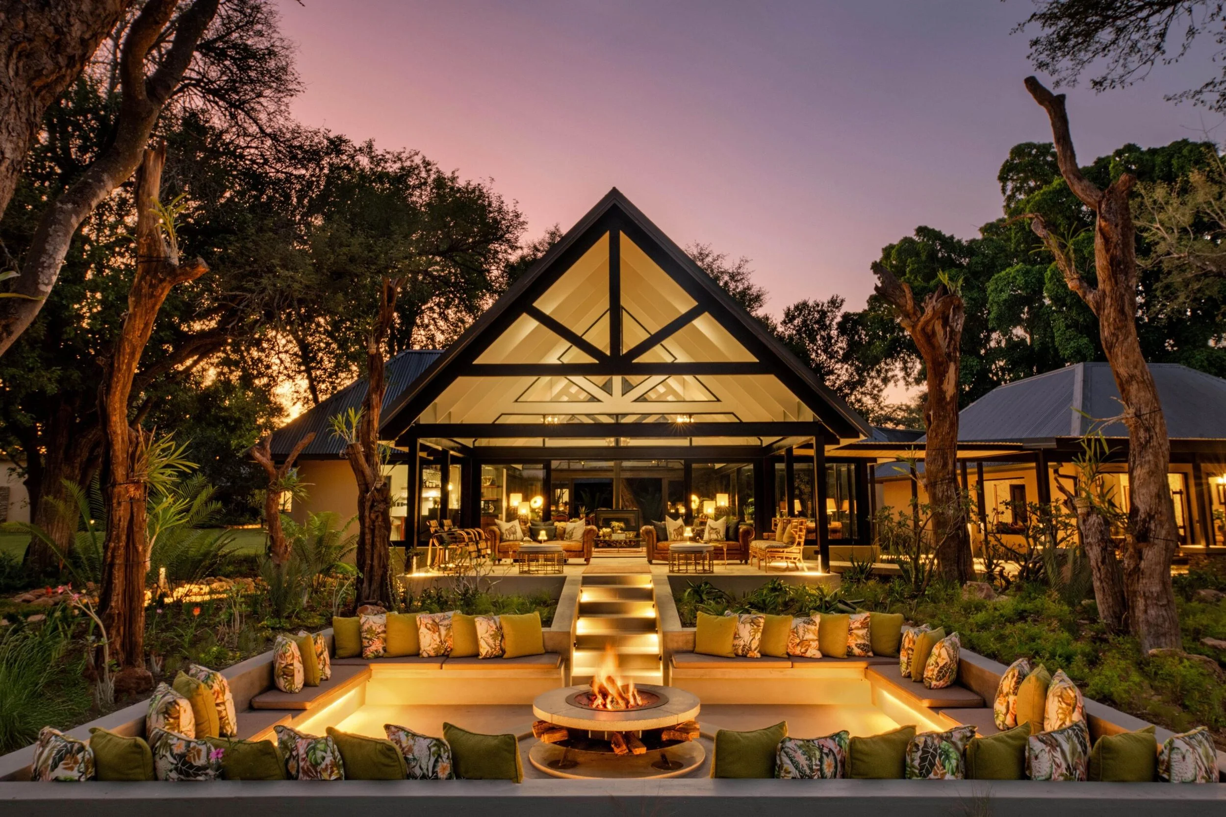Outdoor luxury outdoor living space at sunset with a fire pit surrounded by cushions, a modern house with large glass windows, and trees in the background.