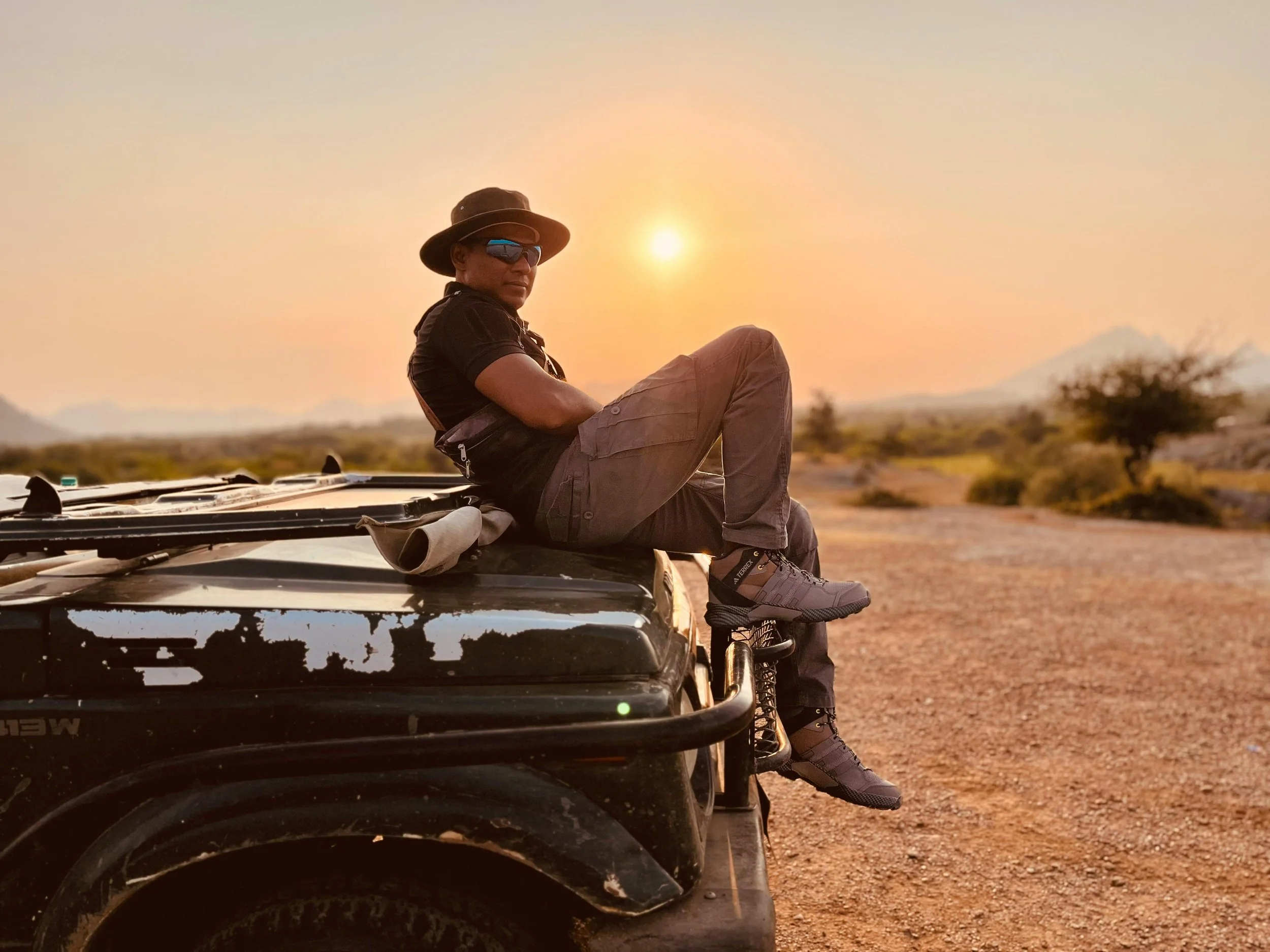Man wearing safari clothing seated on a safari vehicle, surrounded by a natural landscape