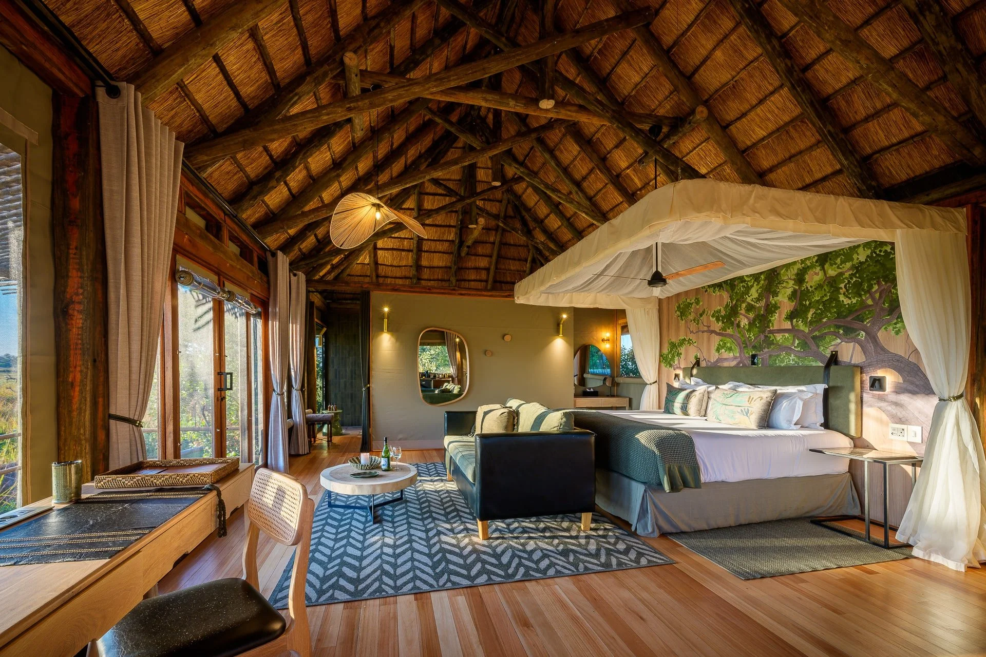 A luxurious bedroom with a thatched roof, large windows with curtains, a king-sized bed with canopy and curtains, a sitting area with a sofa, rug, and coffee table, and a wall painting of a large tree.
