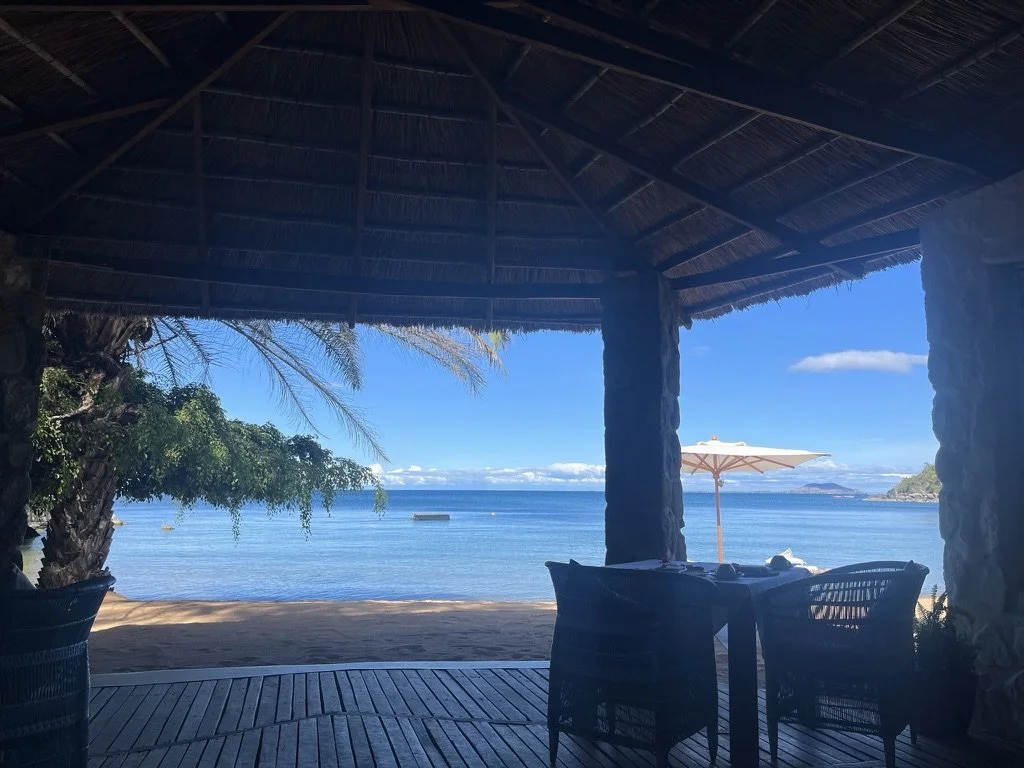 View of the ocean from a shaded beachside patio with tables and chairs, an umbrella, and tropical trees in the background.