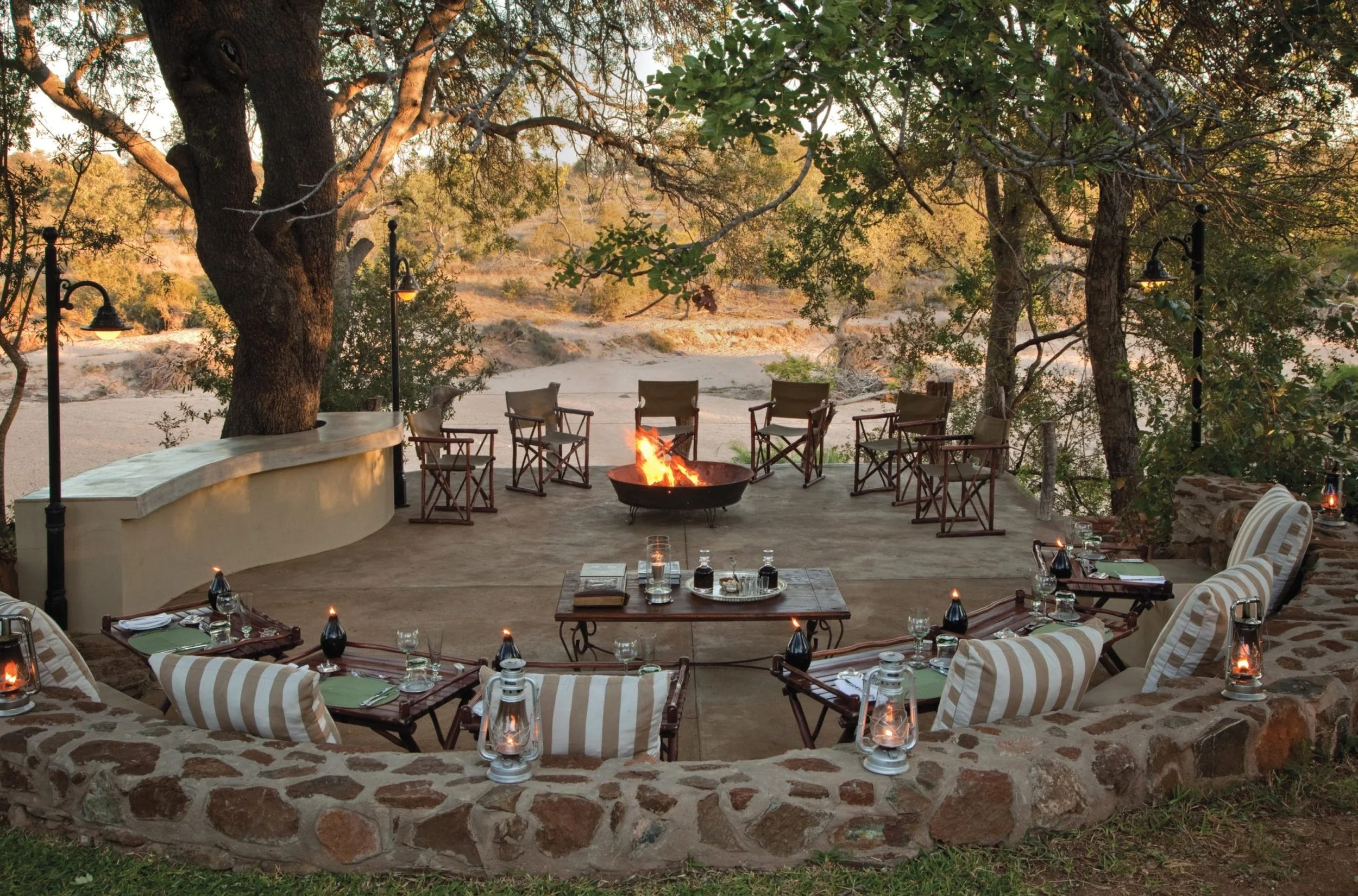 Outdoor dining area with a fire pit, surrounded by chairs and a stone wall, set under large trees at sunset.