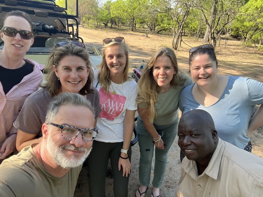 Group of seven people outdoors taking a selfie with trees and a vehicle in the background.