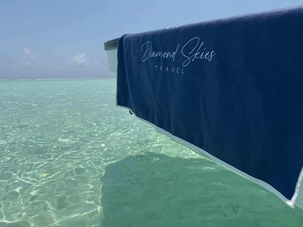A blue beach umbrella with white text reading 'Diamond Skies Travel' floating on clear turquoise water under a partly cloudy sky.