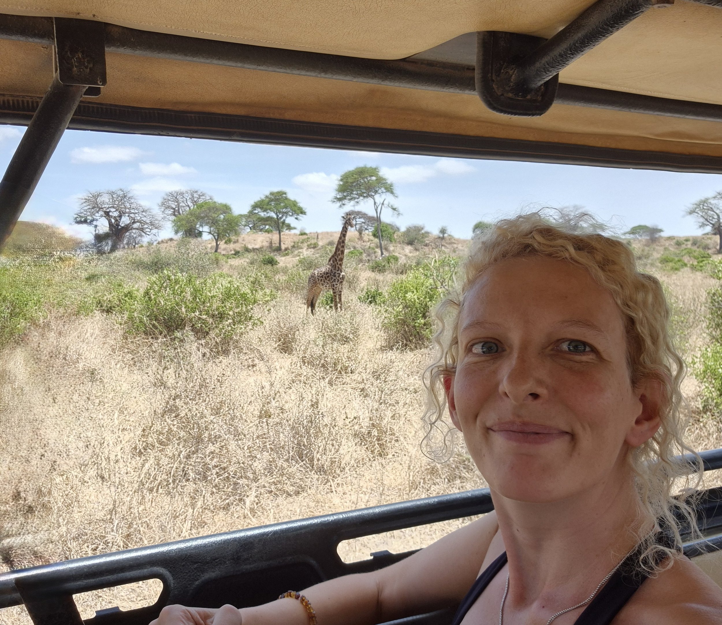 A woman with curly blonde hair smiling at the camera inside a safari vehicle. In the background, a giraffe is standing among trees and bushes in the savannah landscape under a partly cloudy sky.