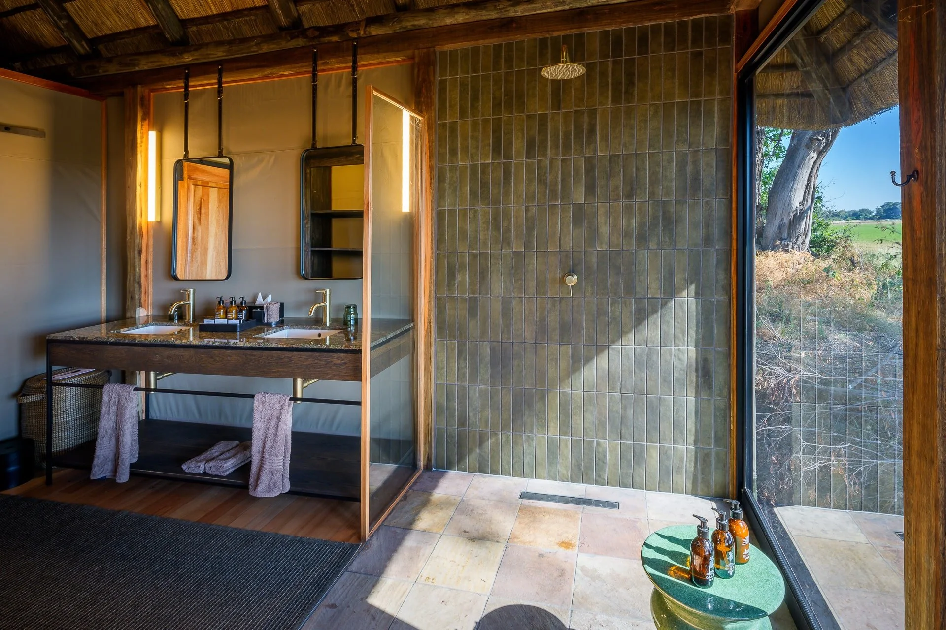 Modern bathroom with dual sinks, wooden accents, mirrors, an outdoor shower area with green tile wall, and a large window showing an outdoor landscape with a tree and field.