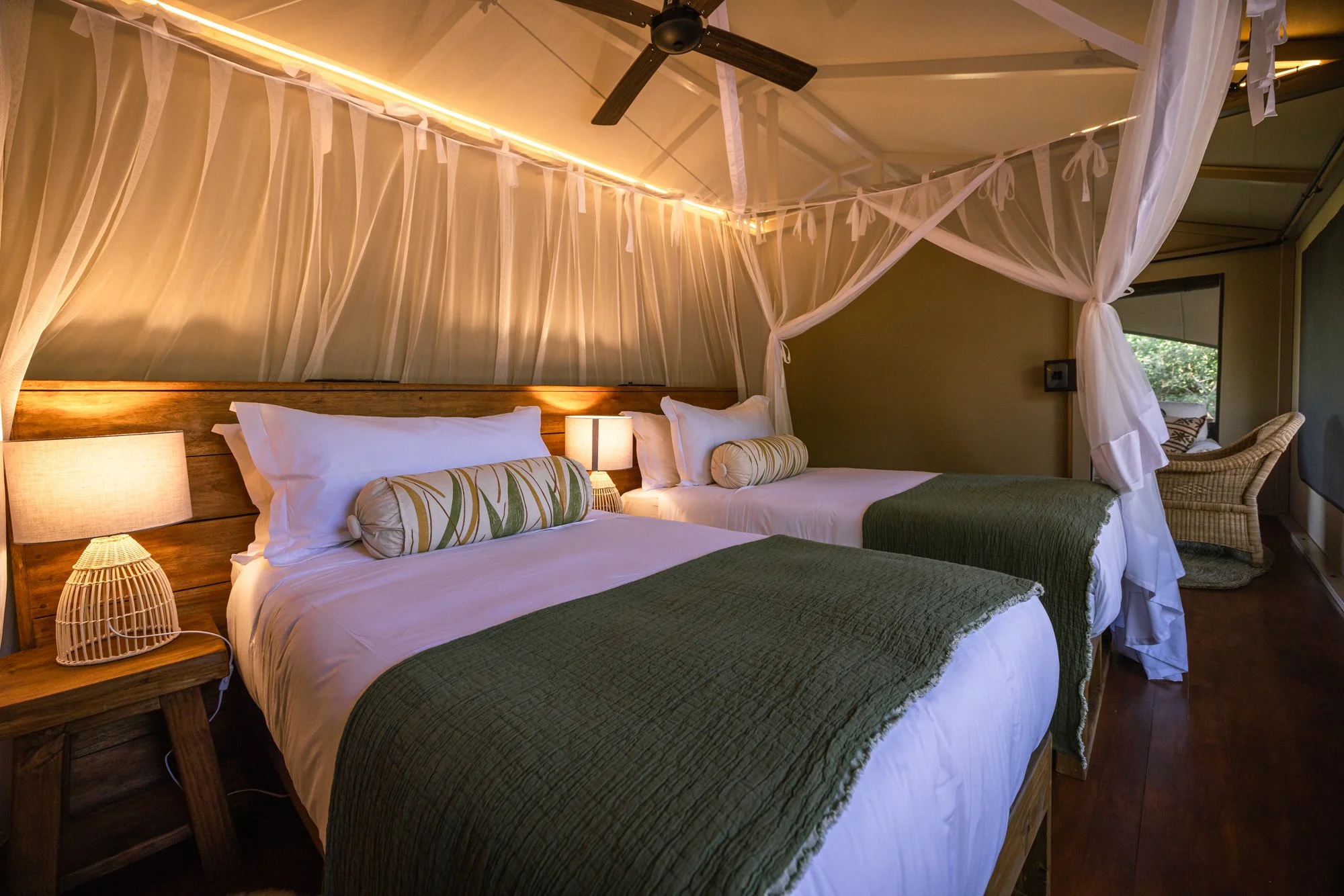 A rustic bedroom featuring two large beds with wooden headboards, white linens, and green bed runners and pillows. A nightstand with a woven wicker lamp is between the beds. The room is decorated with a canopy of sheer white curtains and soft lightin