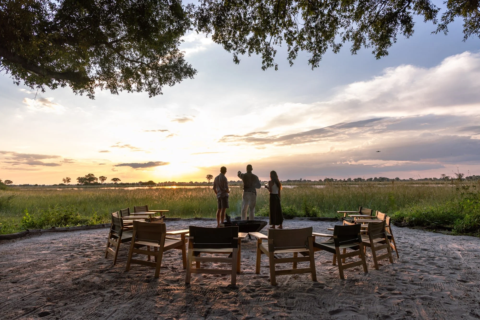 Three people stand around a small firepit on sandy ground, encircled by empty wooden chairs, during sunset near a grassy wetland with a river in the background.