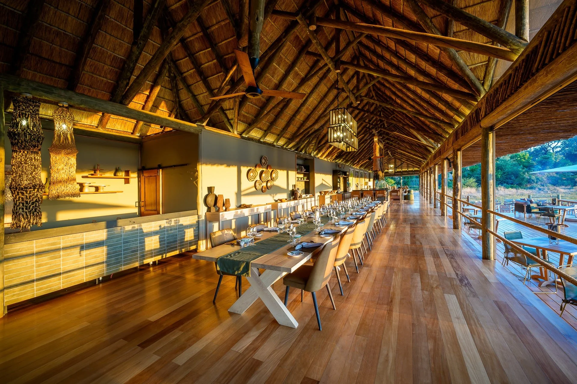 A spacious dining area with a long table set with glassware and napkins, inside a structure with a high thatched roof and large open windows view of outdoor seating area with trees.