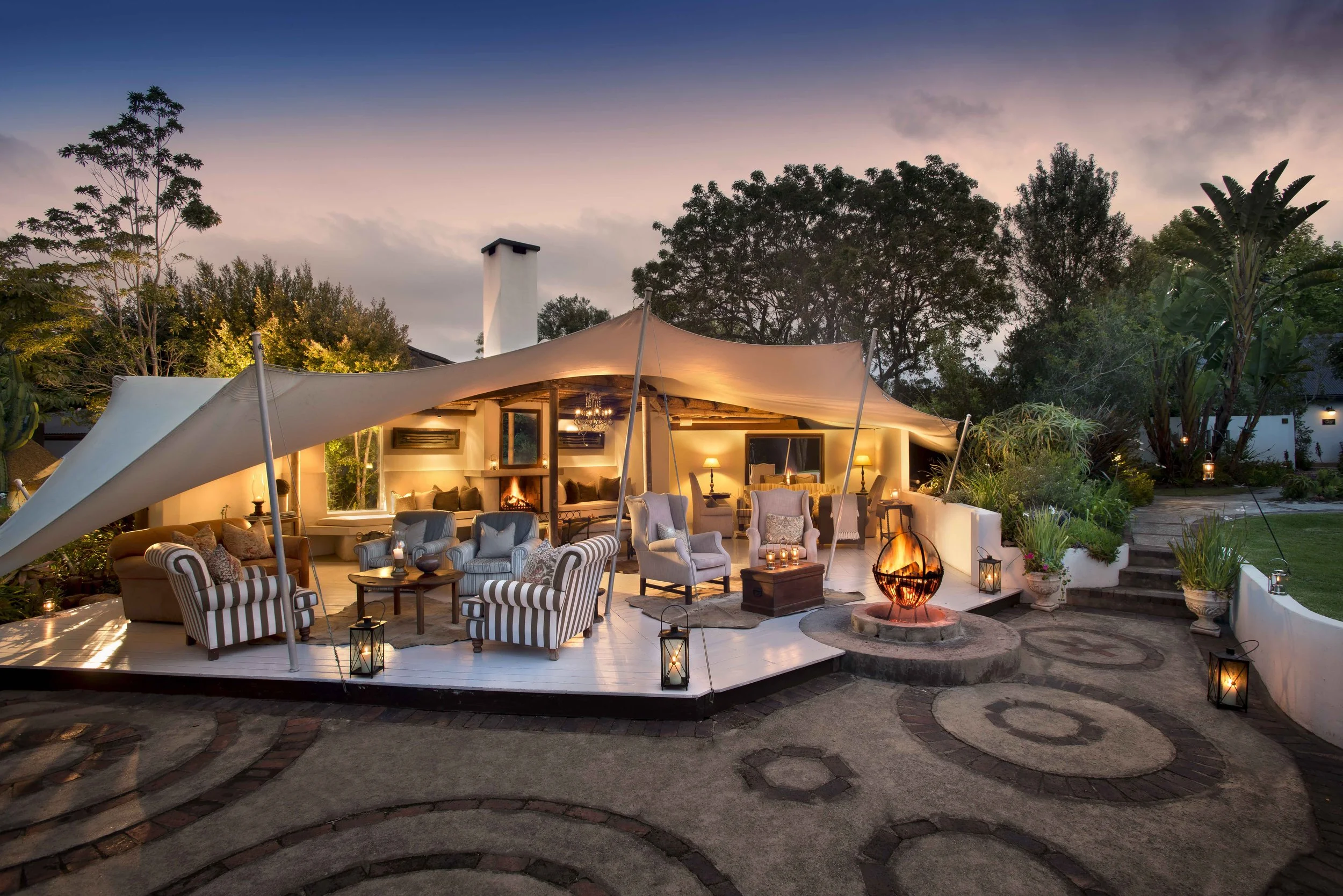 An outdoor patio with a white deck, furnished with striped and patterned armchairs and small tables, illuminated by lanterns and lights, under a canopy at dusk, with trees and a fire feature in the background.