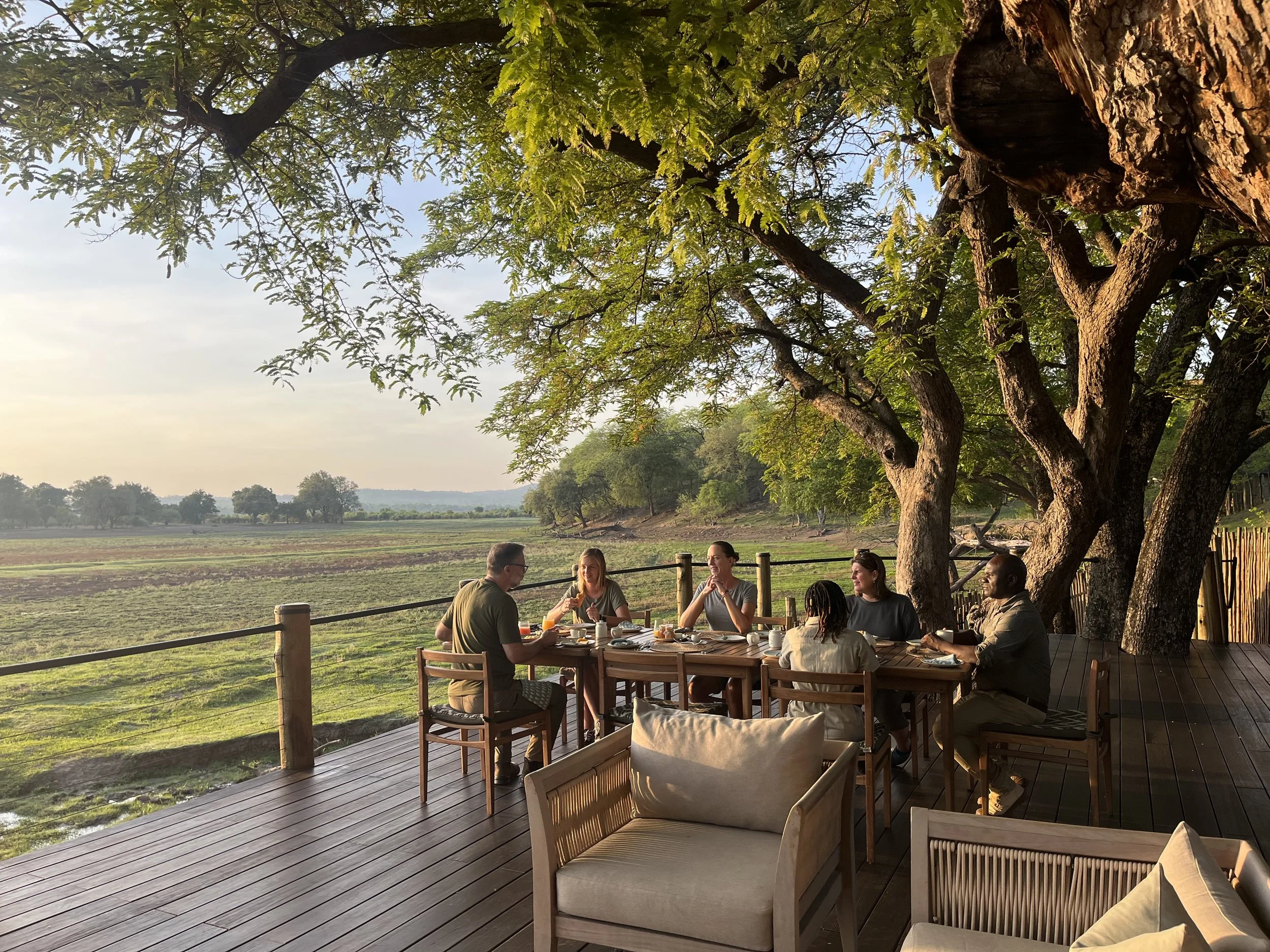 People dining outdoors on a wooden deck under large trees, overlooking a vast open landscape during sunset.