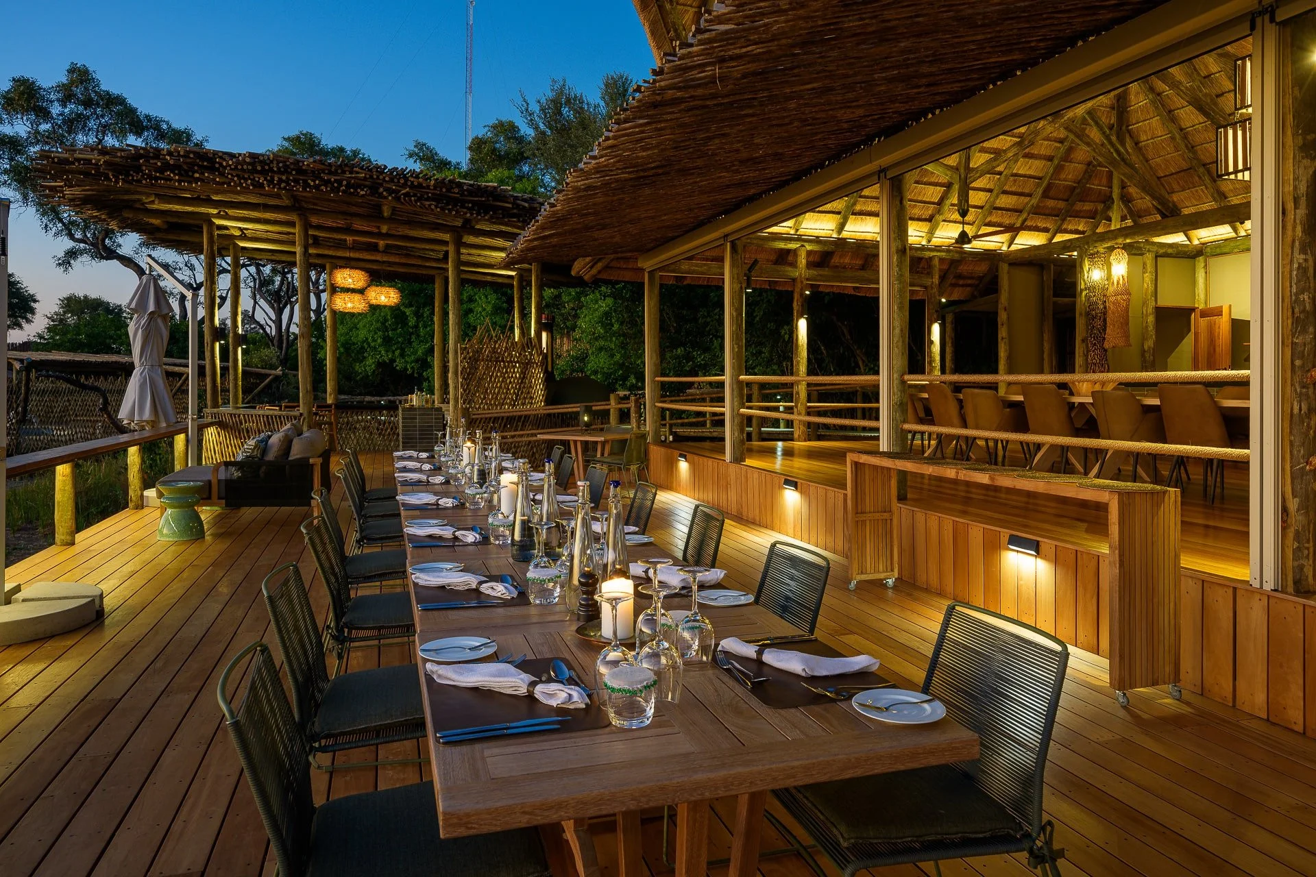 Upscale outdoor dining area with a long wooden table set with wine glasses, plates, and napkins, under a thatched roof structure, illuminated by soft lighting during evening, with trees in the background.
