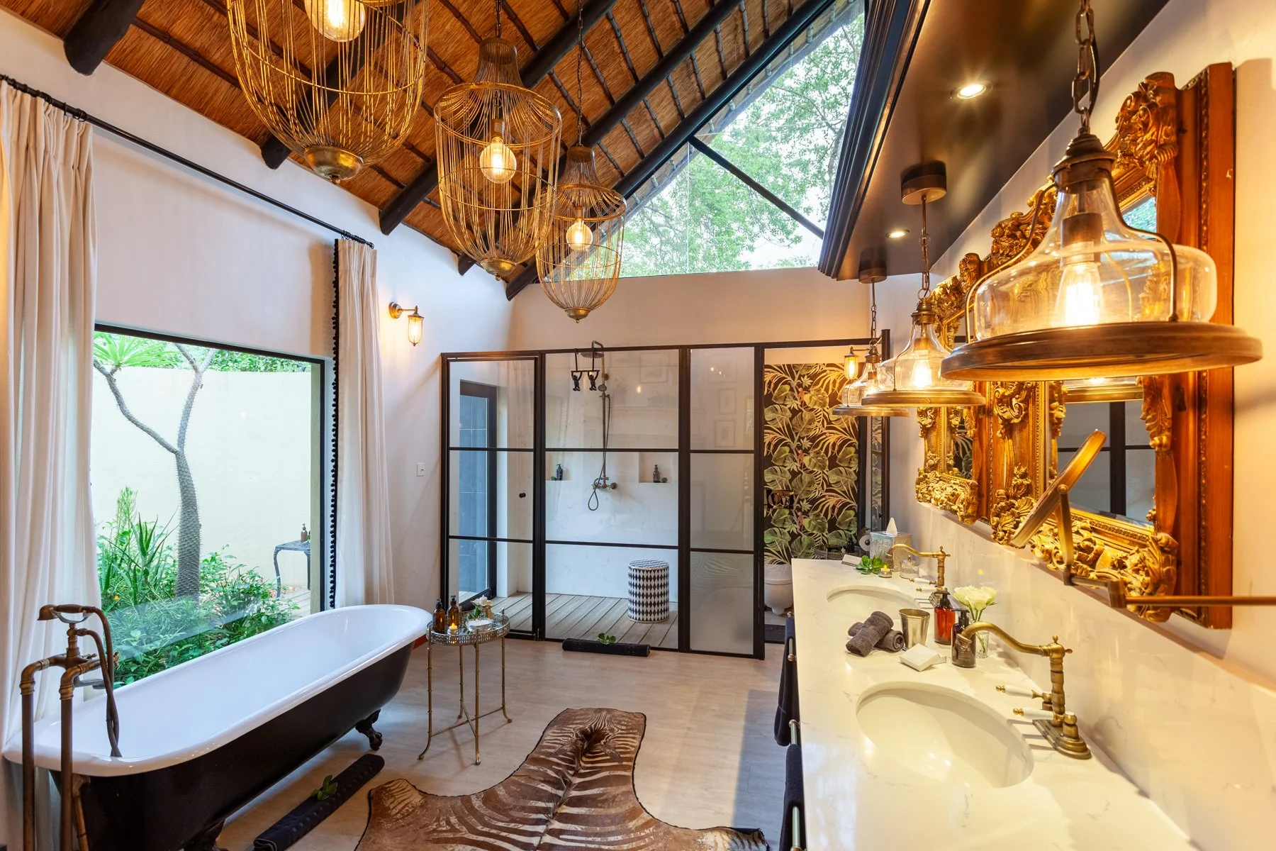 Luxurious bathroom with a freestanding bathtub, large window showing greenery outside, a walk-in shower with glass enclosure, and ornate gold-framed mirrors above a marble countertop with two sinks. Elegant lighting fixtures hang from the ceiling, an